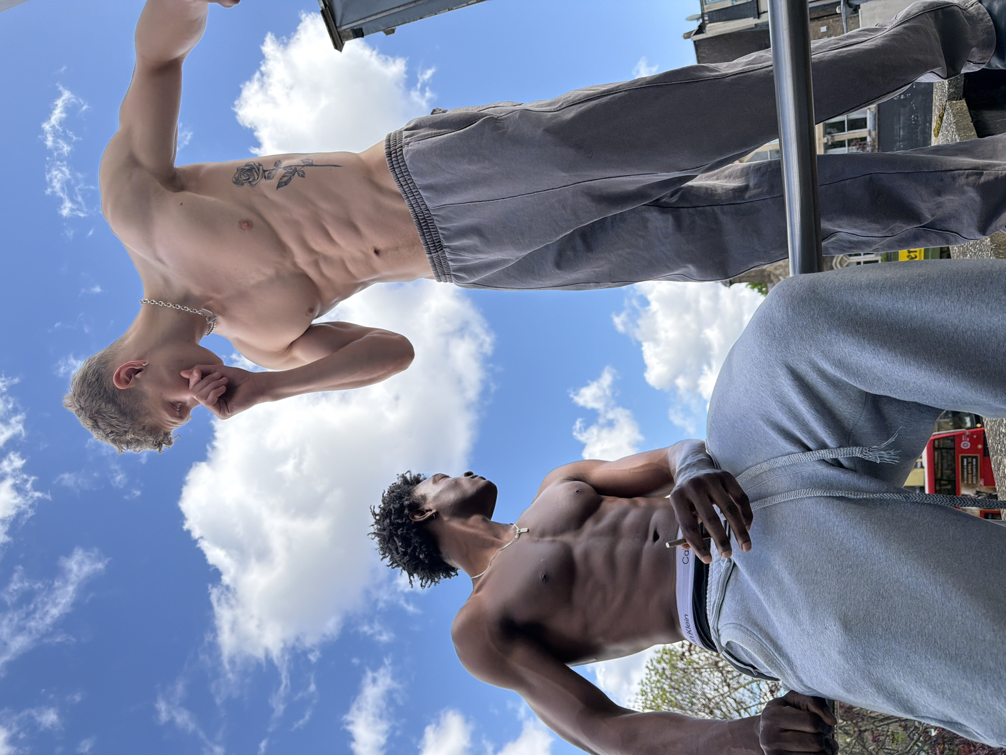 Two men are positioned on a rooftop against a sky with scattered clouds. The man on the upper left has fair skin and short hair, wearing a silver chain and grey sweatpants. He has a rose tattoo on his left rib cage. The man on the lower right has dark skin and short curly hair, wearing a silver chain, grey sweatpants, and Calvin Klein underwear. He is holding a cigarette in his left hand. The background includes building facades and a portion of a red double-decker bus, suggesting an urban location, likely in London, United Kingdom.