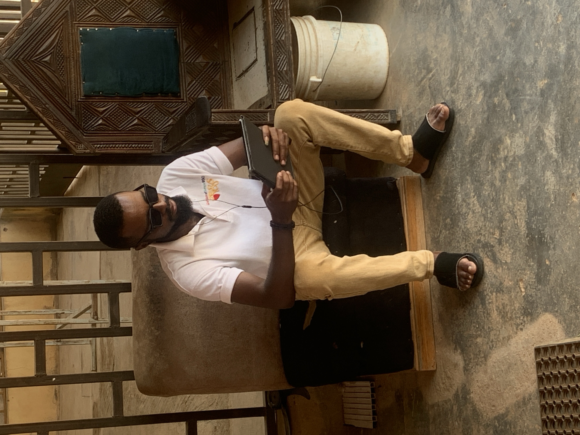 A man with dark skin, a beard, and sunglasses is sitting on a wooden chair and appears to be looking at a tablet. He is wearing a white short-sleeved shirt and tan pants. He is also wearing black sandals. The scene appears to be outdoors, with a textured ground and a wooden structure in the background. There is a white bucket in the background as well. The lighting suggests it could be daytime. The overall mood appears relaxed and casual. Some text is visible on the man's shirt, but it is difficult to read. The location is identified as Jos, Nigeria.