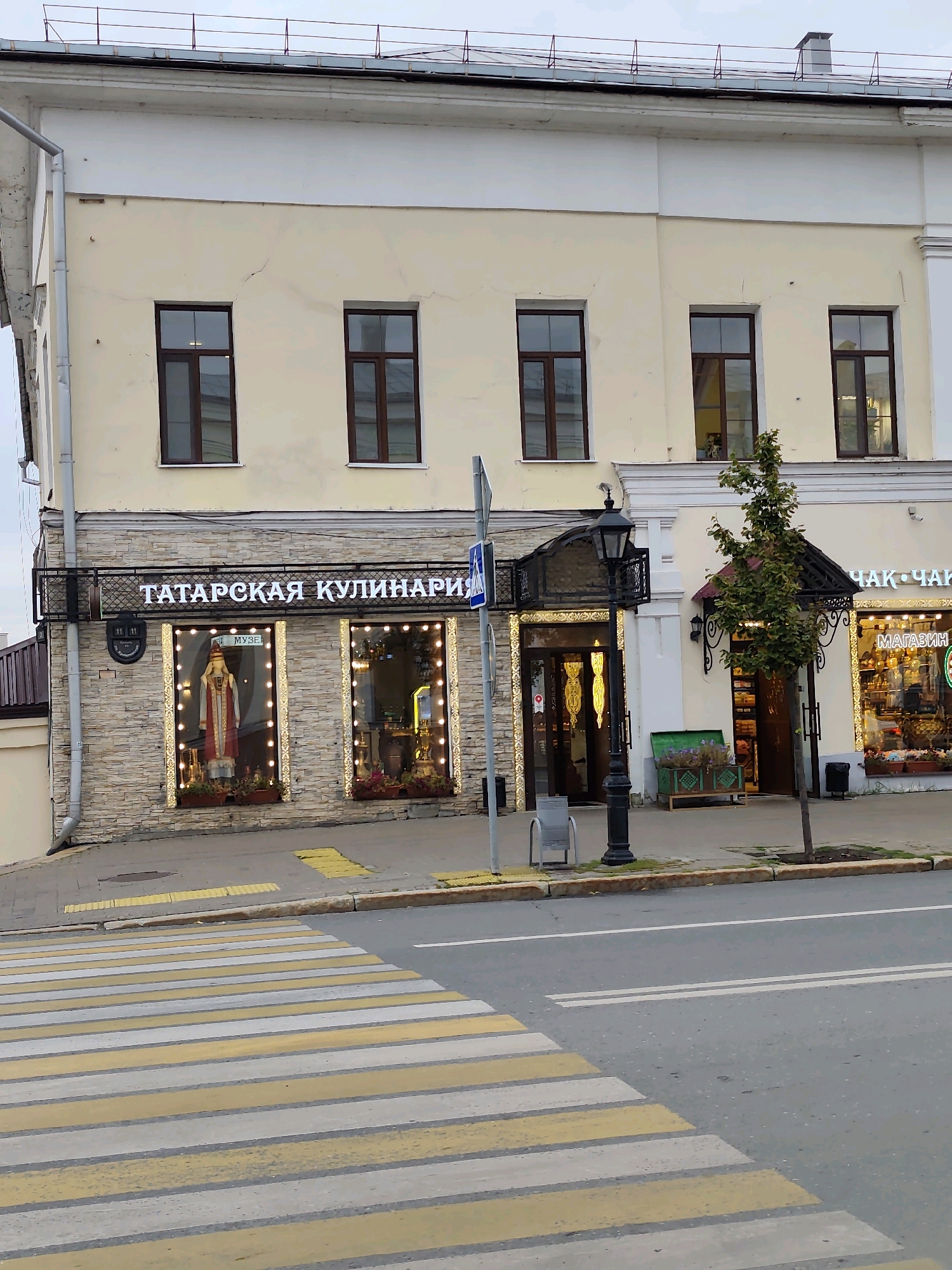 The image captures a street-level view of a building in Kazan, Russia, likely a business establishment. In the foreground, a pedestrian crosswalk with yellow and white stripes dominates the lower portion of the frame, indicating an intersection or a designated crossing area. The building itself is a two-story structure. The lower facade is clad in light-colored stone or brick, featuring a prominent sign that reads "ТАТАРСКАЯ КУЛИНАРИЯ" (Tatar Cuisine) in illuminated white letters. Beneath the sign, two large display windows are visible. The left window showcases a mannequin dressed in traditional attire, with decorative lights framing the window. The right window also appears to be a display, possibly for goods or more decor, also outlined with lights.
To the right of the "Tatar Cuisine" establishment, another storefront is partially visible, bearing the text "ЧАК-ЧАК" (Chak-Chak) and "МАГАЗИН" (Shop). A small tree grows on the sidewalk in front of this section of the building. A traditional black street lamp stands between the two main storefronts. Several trash receptacles are present, along with a bench-like structure with decorative greenery. The upper floor of the building features several uniformly spaced windows with dark frames, set against a light yellow wall. The sky is overcast, suggesting a cloudy day. The scene appears to be part of a city street with sidewalks and a paved road. There are no people clearly visible in the image.