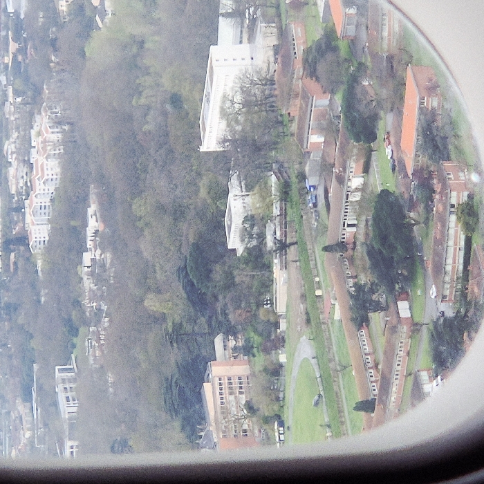 This is an aerial photograph captured from an aircraft, depicting a section of Blagnac, France, under an overcast sky. The view is framed by the aircraft window on the left and bottom, presenting a downward perspective over an urbanized landscape characterized by a mix of institutional, commercial, and residential structures interspersed with significant green spaces.
In the immediate foreground, a cluster of single-story, elongated buildings with red-tiled roofs is visible. These structures, primarily light brown or brick-colored, form several interconnected units, with internal courtyards or open areas containing sparse trees and paved surfaces. Multiple vehicles, including white, red, and dark-colored cars, are parked in these areas. Adjacent to these buildings, a grassy field with a curving dirt path is present, and one dark vehicle is parked on the path. A fence line traverses horizontally across the middle of the lower half of the frame.
Further into the midground, larger multi-story buildings stand out. On the left, a tall, multi-windowed building with an orange-brown façade is prominent. Towards the center-right, two large, light-colored (white or pale grey) multi-story buildings are visible, one taller and more prominent than the other, surrounded by trees. These buildings are situated within an extensive area of deciduous trees, many appearing bare or with minimal foliage.
In the background, beyond the immediate cluster of buildings and trees, more residential areas are discernible. These include multi-story apartment complexes with white facades and red roofs, and some modern-style buildings with horizontal window bands, all nestled amidst further wooded hills and urban development that recedes into the hazy distance. No individuals or specific actions are identifiable within the frame due to the aerial perspective and distance.