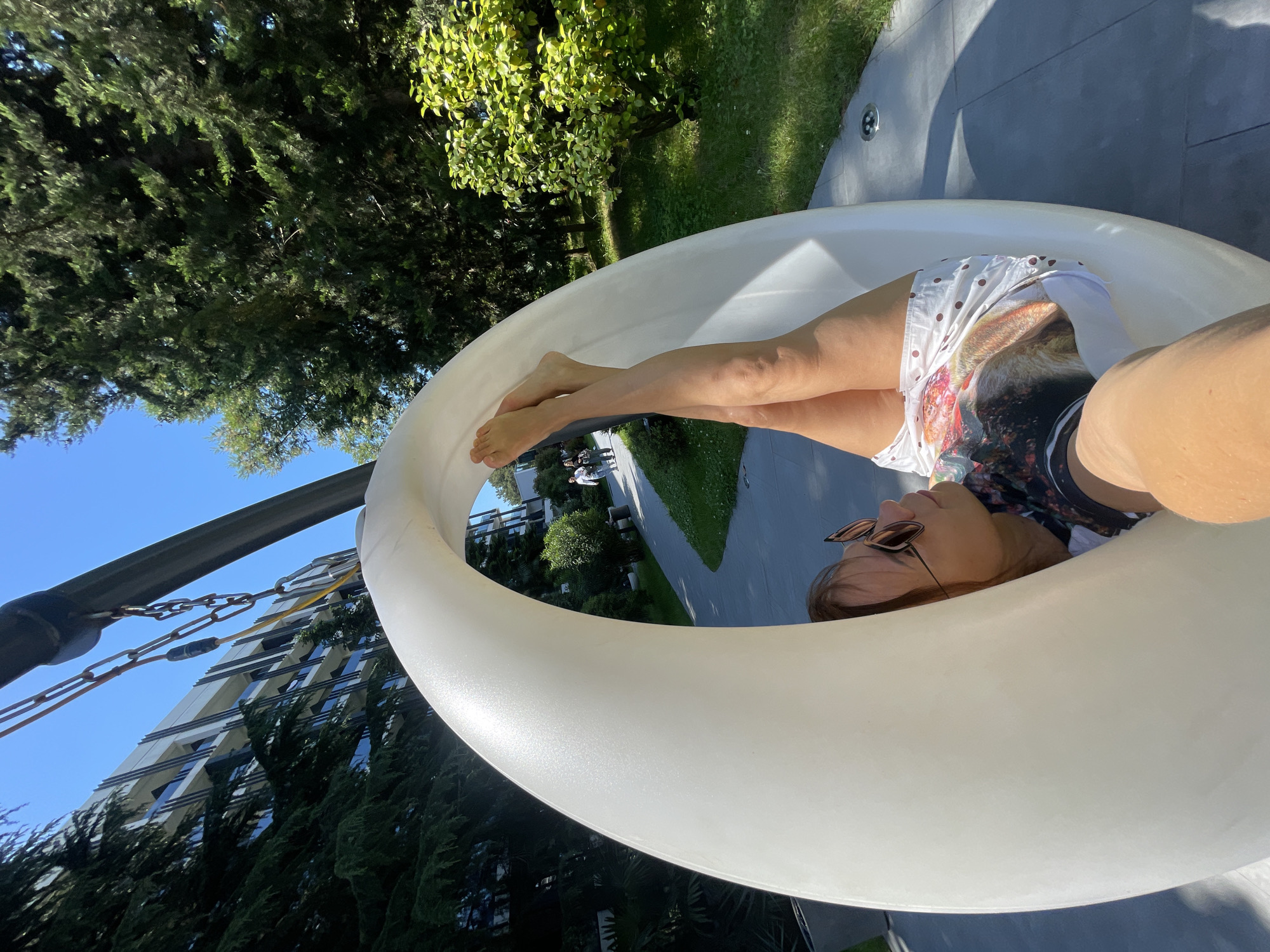 A woman is lying on her back in a large, white, oval-shaped hanging chair. Her legs are extended upwards, crossed at the ankles, and are visible above the top edge of the chair. She is wearing white shorts with a polka dot pattern and a t-shirt with a graphic print. She has sunglasses on and is looking to her left. The chair is suspended from a metal frame, with chains visible.
The background shows a sunny outdoor environment. To the left, there is a multi-story building with balconies and glass windows, surrounded by tall, dark green coniferous trees. To the right of the building, a paved pathway winds through a grassy area with more trees and bushes. Two people are walking on the pathway in the distance. The sky is clear and blue. The scene appears to be in a resort or park area, possibly in Sochi, Russia, given the architecture and vegetation.