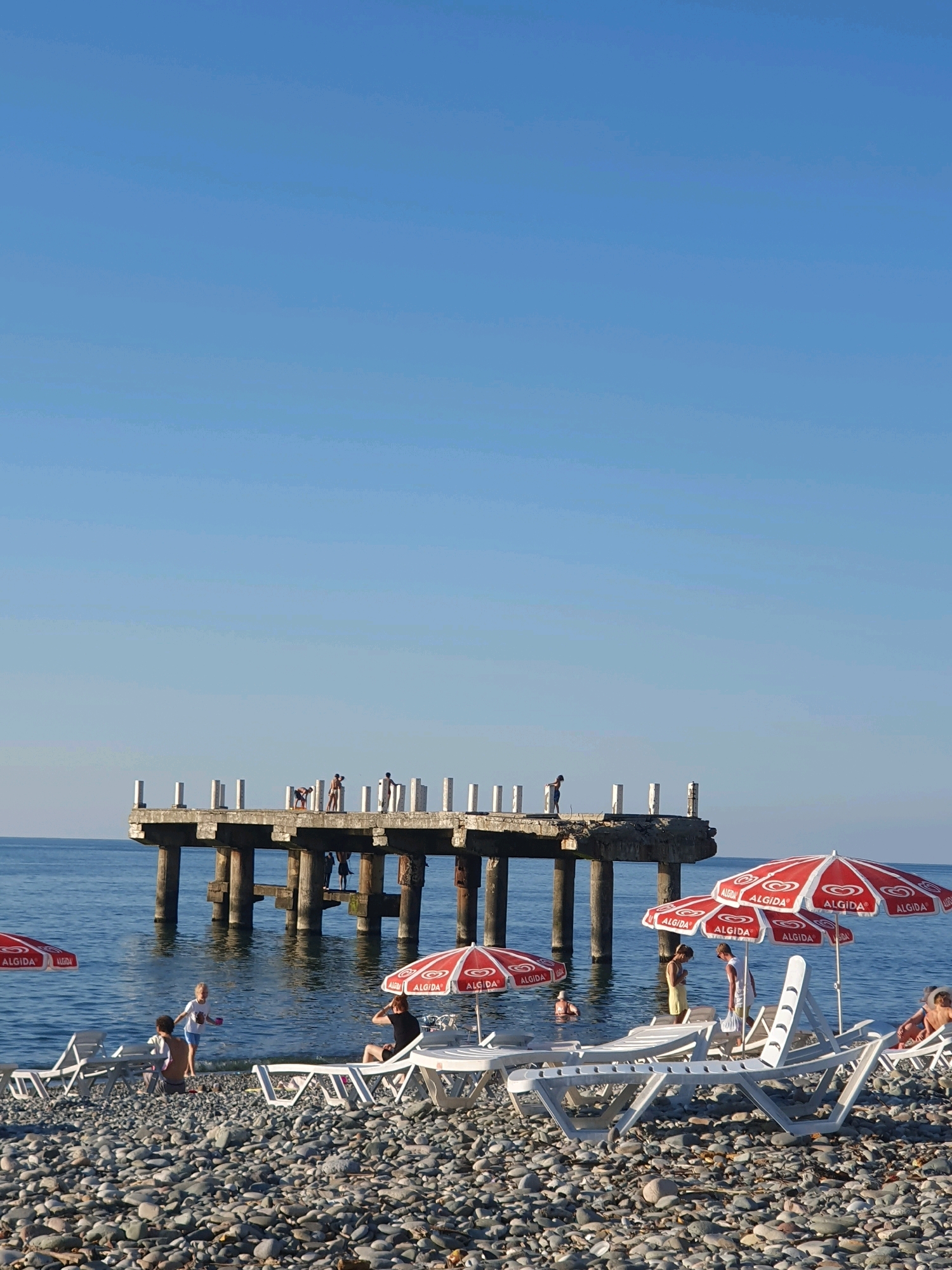 This is a photograph taken at a beach in Batumi, Georgia, with a clear blue sky. In the foreground, a pebbly beach is dotted with white lounge chairs, some occupied by people relaxing. Several red and white umbrellas, branded with "ALGIDA," provide shade. In the midground, people are wading in the calm blue sea. Dominating the background is a weathered concrete pier extending into the water, with white pillars lining its edge. Several figures, appearing to be children or teenagers, are visible on the pier, some standing and one appearing to be in motion, possibly jumping. The overall scene depicts a typical summer day at a coastal resort.