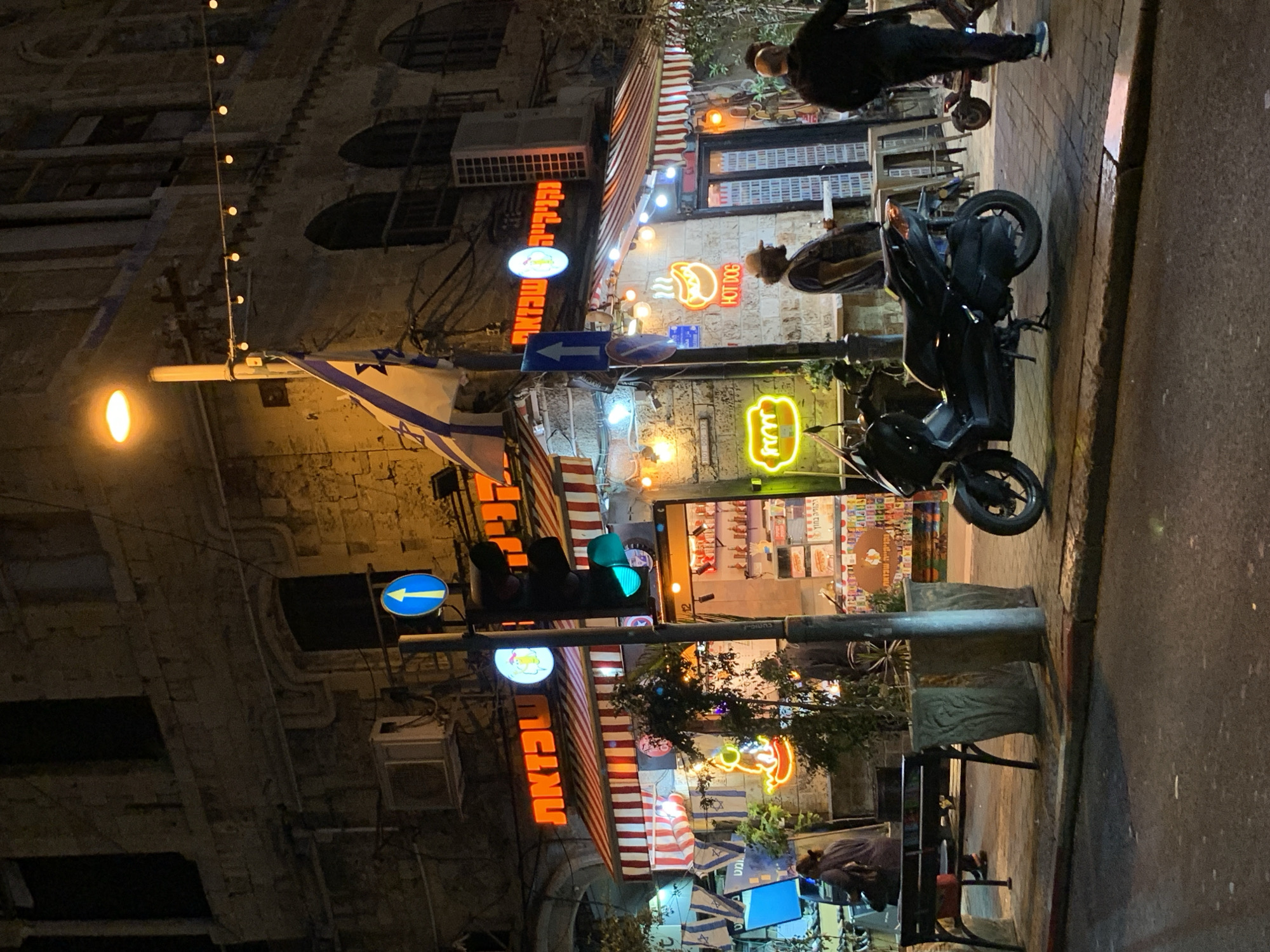 The image depicts a street scene in Tel-Aviv, Israel, at night. A building on the left is constructed of stone. Neon signs advertising food establishments are illuminated along the street. Pedestrians can be seen on the sidewalk. A parked motorcycle is present. There is an Israeli flag present. Streetlights and a traffic light are visible. A blue street sign is present. A bench and planters can be seen on the sidewalk.
