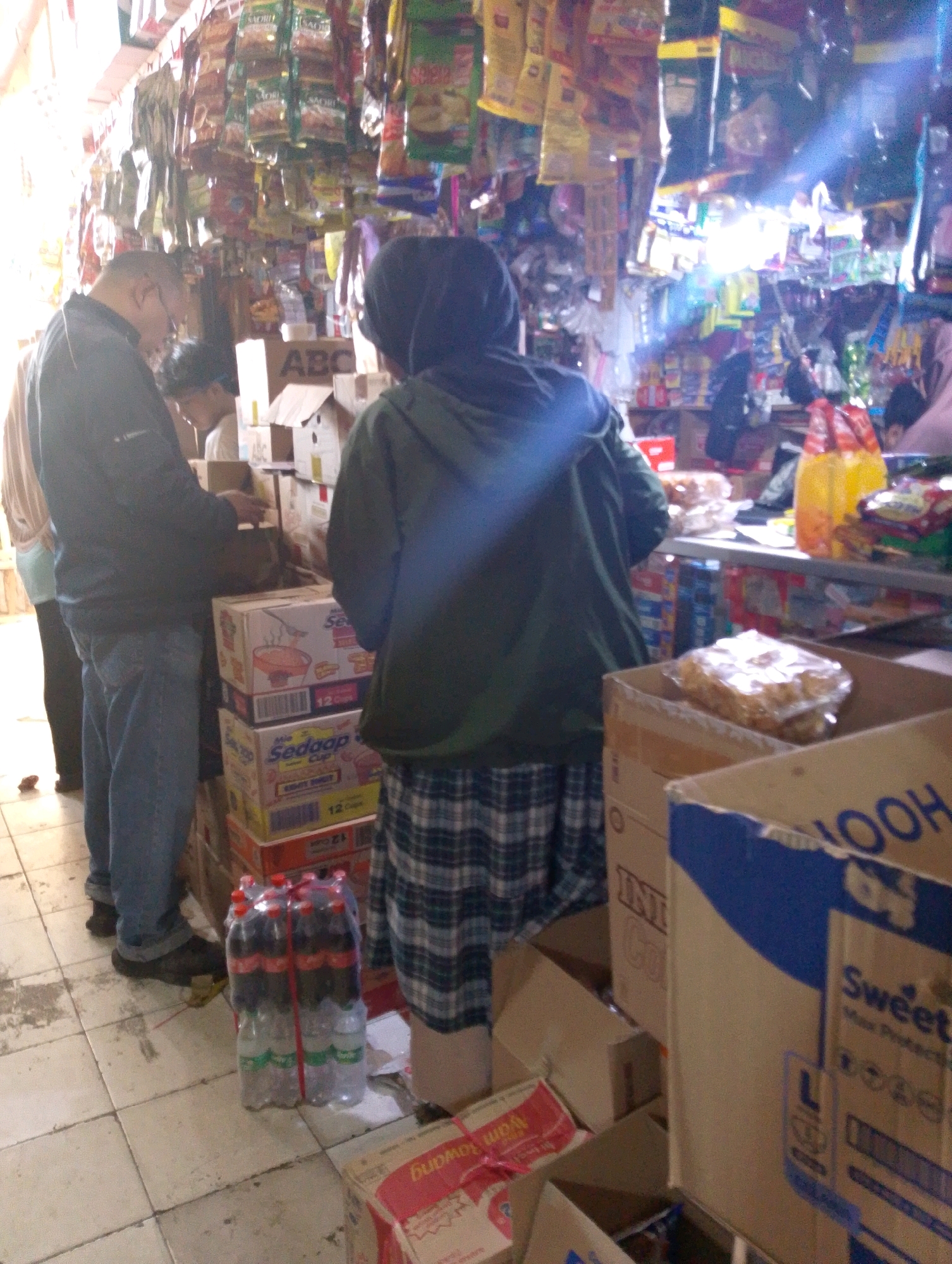 The image depicts the interior of a small, cluttered convenience store in Lembang, Indonesia. The scene is dominated by shelves overflowing with various packaged goods, many of which hang from the ceiling. The lighting appears to be a mix of natural light from an unseen source and artificial lighting within the store, creating some glare.
In the foreground, on the left, a man in a dark jacket and blue jeans is standing. He is facing towards the right of the frame, partially obscured by his jacket. To his right, a person wearing a dark hooded garment is standing, facing away from the camera and towards the interior of the store. Their lower body is clad in a plaid skirt.
Numerous cardboard boxes are stacked on the floor and on shelves throughout the store. Some boxes are open, revealing their contents. One prominent stack of boxes in the center features branded packaging for what appears to be instant noodles or similar food products, with visible text like "Sedap."
A six-pack of plastic bottles, likely beverages, is placed on the floor in front of a stack of boxes. The bottles are clear with colored liquid and are secured with red ties.
In the background, more shelves are visible, filled with a dense array of products, including snacks, drinks, and other household items. A yellow plastic bag and a red plastic bag are also visible on a shelf in the background, suggesting merchandise or shopping bags. The overall impression is one of a busy, well-stocked local store.