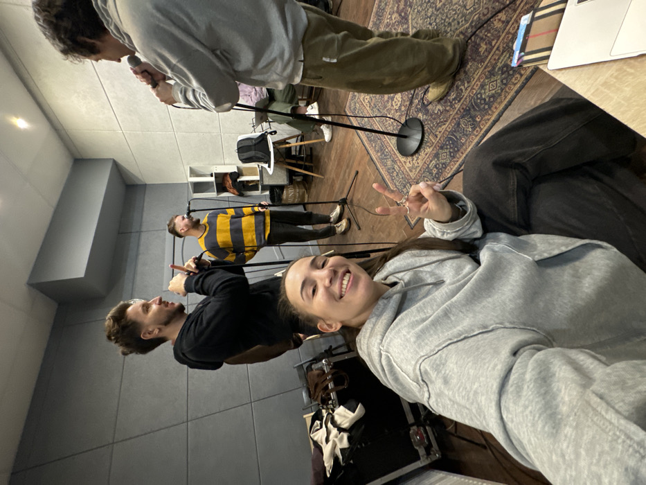 The image depicts a casual gathering of five individuals within a recording studio in Moscow, Russia. In the foreground, a female with long dark hair, wearing a grey hooded sweatshirt and dark pants, is taking a selfie with a mobile device, smiling directly at the camera while making a peace sign with her right hand.
Behind her, three males are actively engaged with microphones. To the left, a male with a beard and dark hair, dressed in a black hooded sweatshirt, holds a microphone on a stand and gestures with his left index finger. In the center, another bearded male, wearing a yellow and dark horizontal striped sweater, stands facing towards the left, holding a microphone on a stand. To the right, a third male, wearing a light grey hooded sweatshirt and olive-green pants, holds a microphone near his mouth, facing towards the other two men. A fourth individual is partially visible, seated in the background.
The studio environment features grey acoustic foam panels covering the walls. The floor is dark wood, with a patterned rug visible in the right foreground. Equipment includes multiple microphone stands, a black equipment case on the left, and a small table with a black backpack and a partial view of a laptop on the right. The overall scene suggests a rehearsal or recording session.