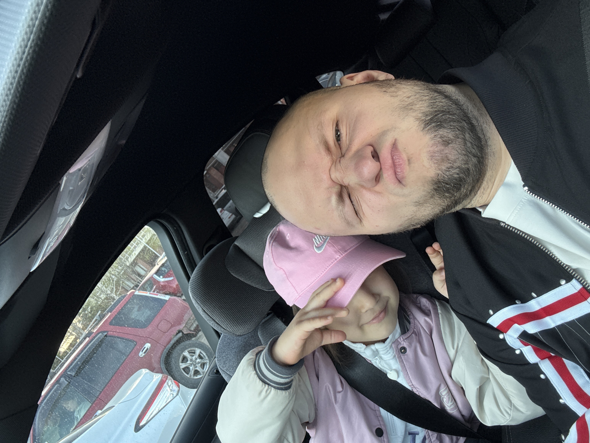 A man and a young girl are in a car. The man is bald with a beard and is wearing a black jacket with white, red and black stripes. The girl is wearing a pink Nike baseball cap and a light pink jacket. She is in a car seat and wearing a seatbelt. The background shows other cars and an urban setting, possibly in Krasnoyarsk, Russia.