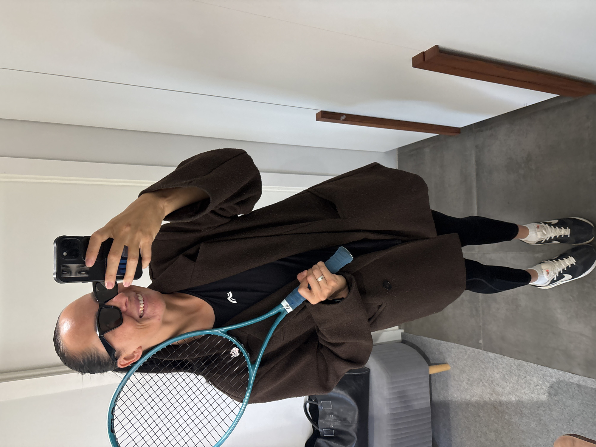 A person is taking a selfie indoors, holding a blue tennis racket and a smartphone. The person is wearing black leggings, black sneakers with white detailing, a black t-shirt with a white logo, a brown coat, and sunglasses. The environment includes a grey floor, white walls, and what appears to be a wooden beam on the ceiling. There is also a grey chair or ottoman visible in the bottom right corner. The location in the city of Chaikovsky, Russia is not discernible from the image.