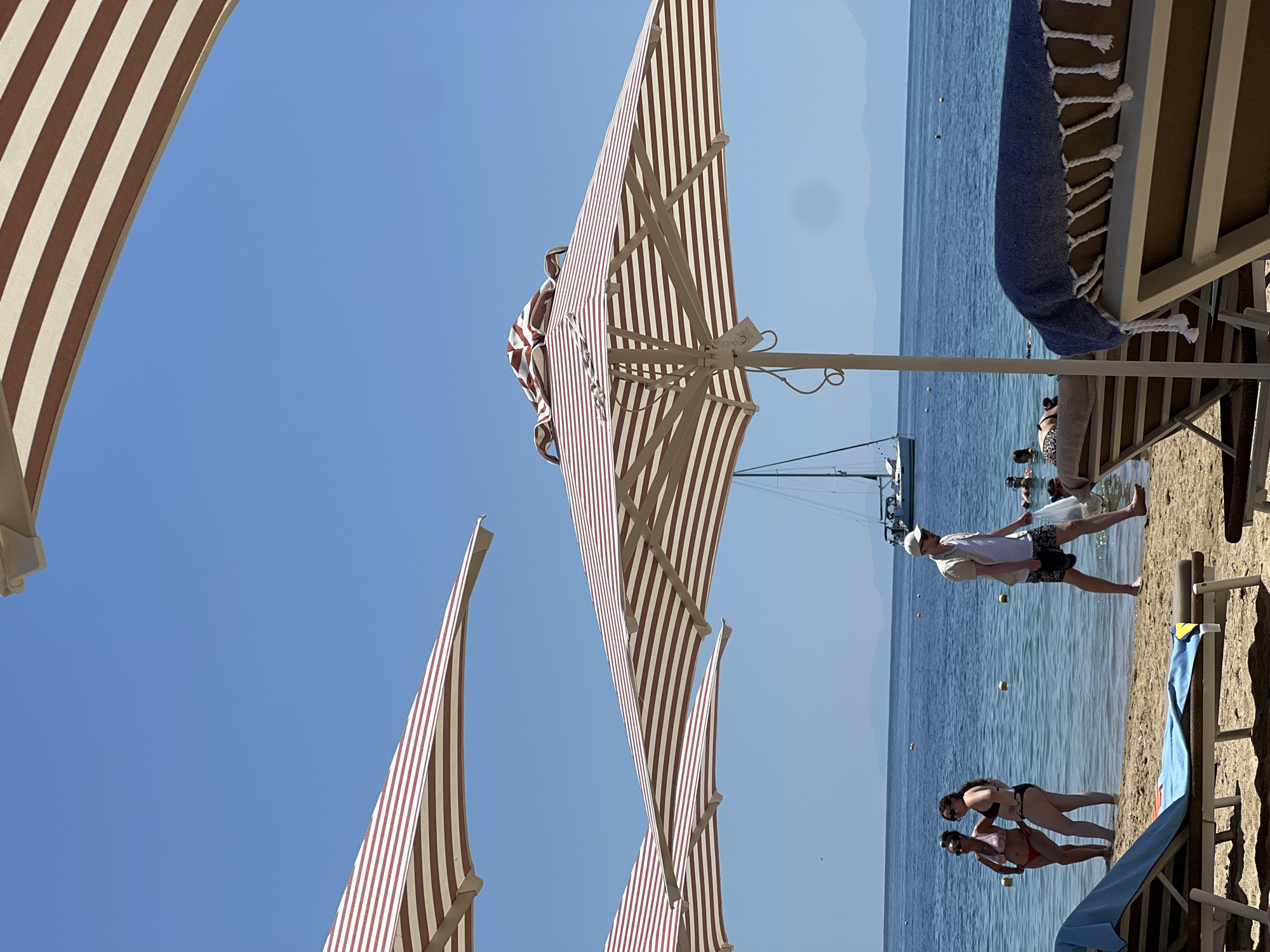 The image depicts a sunny day at a beach in Troulos, Greece. Several beach umbrellas with red and white stripes are set up on the sand. In the foreground, two lounge chairs are visible, one with a blue towel draped over it. The turquoise sea is calm and clear, with a few people swimming and a sailboat in the distance. Two women are standing in the shallow water, and a man is walking on the beach carrying a plastic bag. The sky is a clear blue, and mountains can be seen in the background.