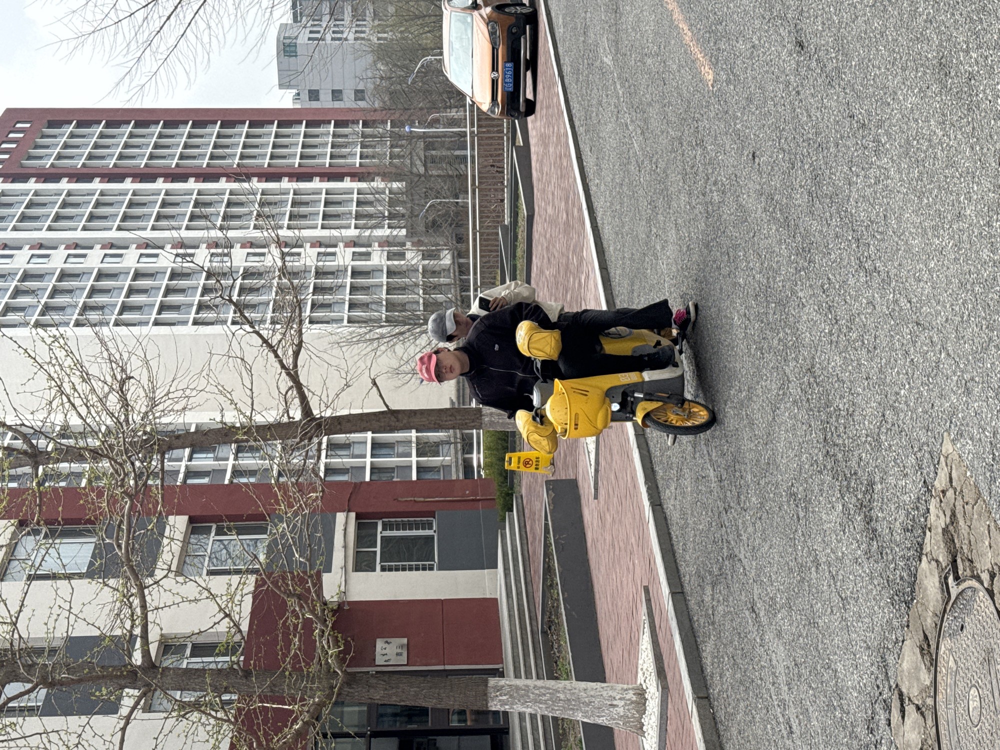 The image depicts an outdoor street scene in Zhongtun, China, on what appears to be an overcast day, likely in late winter or early spring given the bare branches of the trees.
In the foreground, two individuals are seated on a bright yellow electric scooter, positioned on the edge of a brick-patterned sidewalk next to a grey asphalt road. The person in front, wearing a black jacket and a pink baseball cap, looks directly at the viewer. Behind them, another person is partially visible, wearing a lighter top and a grey cap, holding a mobile phone. A small yellow sign, displaying a red circle with a diagonal line (a universal no-entry or prohibition symbol), is mounted on the front of the scooter.
Further down the road, an orange car is parked, its license plate "京 B989B" clearly visible, indicating it's registered in Beijing. The road features a textured asphalt surface, and a circular metal manhole cover is present in the lower right corner.
The background is dominated by large, multi-story buildings. One prominent structure features horizontal red-brown bands alternating with rows of windows set in white frames against a light-colored facade. Another building, partially obscured by the leafless trees, is visible further back. A sign bearing the Chinese characters "三舍" (Sān Shè), likely indicating "Building Three" or "Third Dormitory," is visible on one of the buildings. The sky is uniformly grey, suggesting a cloudy day.