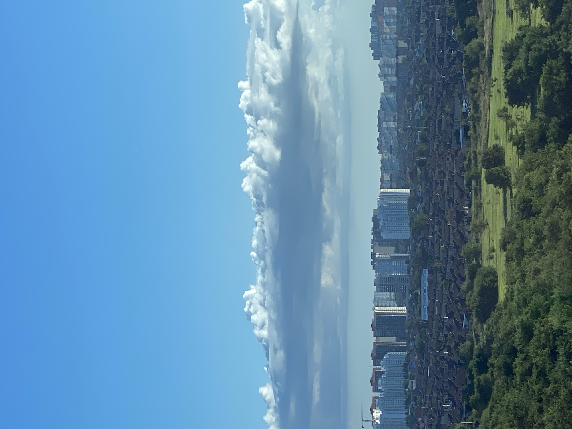 This is a wide shot taken from an elevated position, looking out over a sprawling urban and suburban landscape under a bright blue sky with scattered clouds. In the foreground, lush green trees and vegetation dominate, with a few patches of mowed grass.
As the view moves further back, rows upon rows of red-tiled roofs of houses stretch across the midground, indicating a densely populated residential area. Behind this, a line of taller apartment buildings forms the city skyline. These buildings vary in height and color, with some appearing to be modern high-rises. A construction crane is visible on the far left of this section.
The sky occupies the top half of the image, with a clear blue expanse gradually meeting a band of white and grey cumulus clouds that are spread horizontally across the horizon. The lighting suggests it is daytime, likely morning or afternoon, with the sun illuminating the scene from the side, casting soft shadows. The overall impression is of a vast, developing city with a mix of low-density housing and modern apartment complexes, set against a backdrop of natural greenery and a dynamic sky. This scene is characteristic of the eastern outskirts of Krasnodar, Russia, specifically areas like the "Sunny" (Solnechny) district or nearby developing regions that feature a mix of private housing and new apartment blocks.