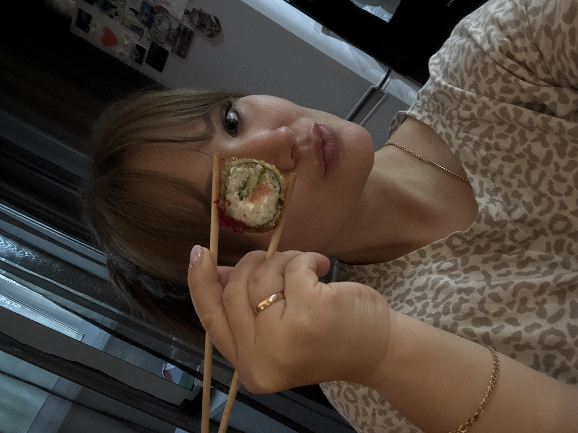The image shows a woman holding a piece of sushi with chopsticks in front of her eye. She is wearing a leopard-print t-shirt and gold jewelry. The background is blurred, but appears to be a domestic setting with curtains and a refrigerator with stickers. There is no indication of the city of Chaikovsky, Russia in the image.