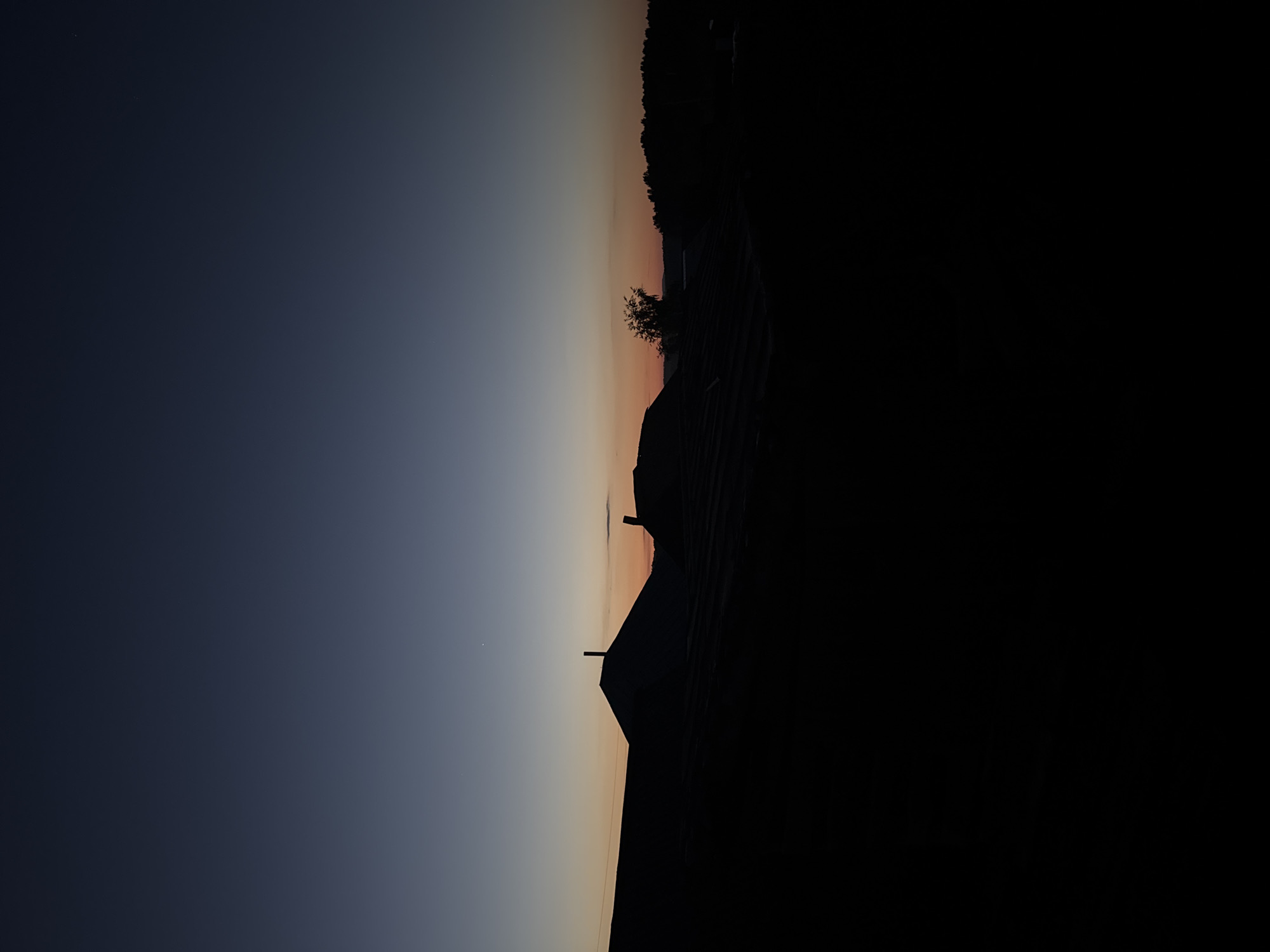 The image shows a silhouetted landscape at dusk. The foreground is dominated by the dark shapes of rooftops, casting long shadows. Beyond the rooftops, the sky transitions from a deep blue at the top to a pale orange and yellow near the horizon, indicating the setting or rising sun. A few faint stars are visible in the upper part of the sky. In the middle ground, silhouetted trees and a single tree with more detail can be seen against the lighter sky. The overall impression is one of quiet and stillness.