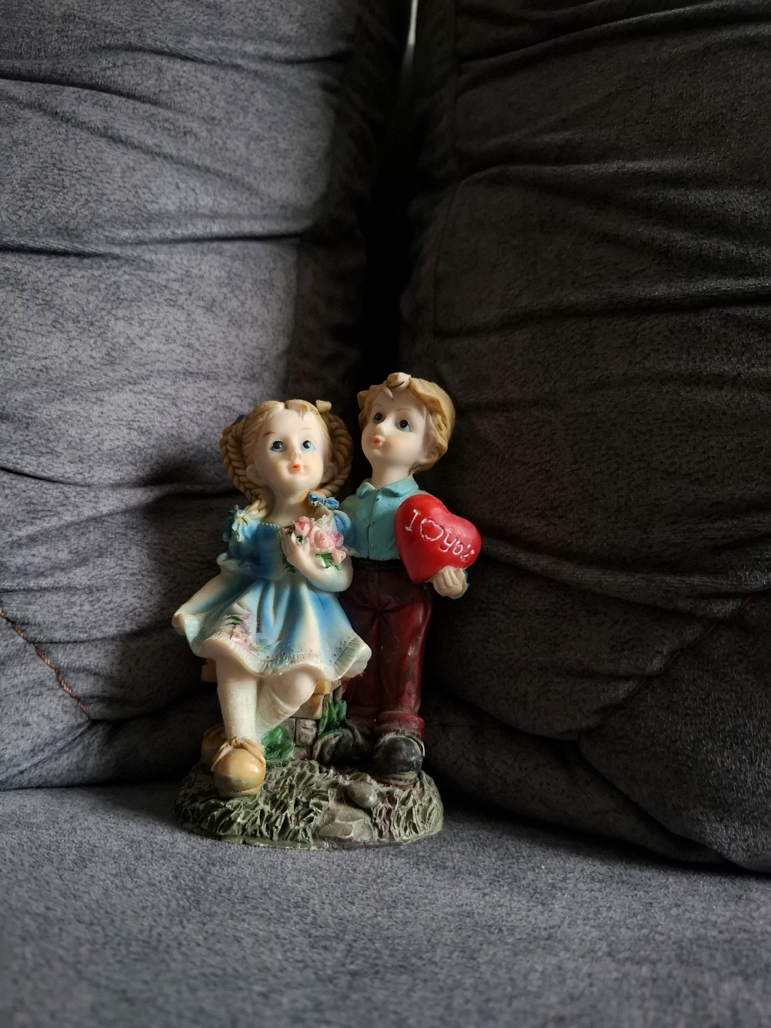 A figurine of a boy and a girl is displayed on a textured gray surface. The boy, on the right, wears a blue shirt and red pants, holding a red heart inscribed with "I love you." The girl, on the left, has blonde hair in pigtails and wears a blue dress. She holds a bouquet of pink flowers. The figurine is placed on a circular base decorated with green grass and rocks, on what appears to be a sofa or upholstered furniture. The background is a dark, heavily shadowed textured fabric. The scene is located in Beyləqan, Azerbaijan.