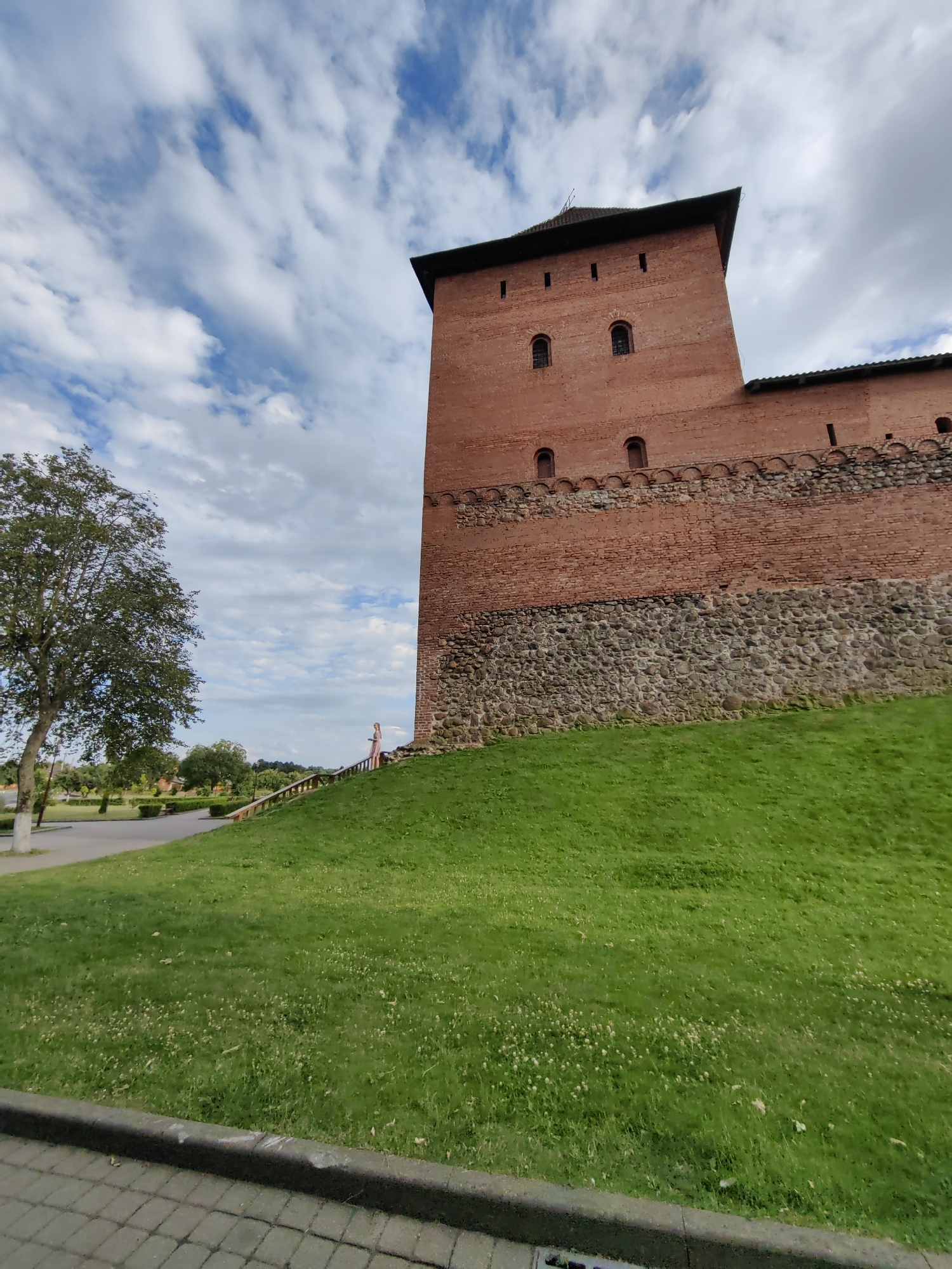 This is a low-angle, wide shot of a brick tower and wall of a historical structure, possibly a castle or fortress. The architecture is ancient, with rough-hewn stone forming the lower portion of the wall and red brick the upper. Small arched windows and defensive openings are visible in the brickwork.
In the foreground, a vibrant green lawn slopes upwards towards the structure. A paved pathway with a curb runs along the bottom edge of the frame. To the left, a mature tree with lush green foliage stands, its trunk painted white at the base. In the middle ground, a wooden fence follows the contour of the grassy slope. A person, appearing to be a woman in a light-colored dress, is visible standing on the fence's walkway, looking towards the tower.
The sky above is a dynamic mix of blue and white clouds, suggesting daytime with possibly some wind. The lighting indicates it is likely late afternoon or early evening due to the shadows cast. The scene evokes a sense of historical grandeur and a peaceful, scenic outdoor setting. No visible text is present in the image.