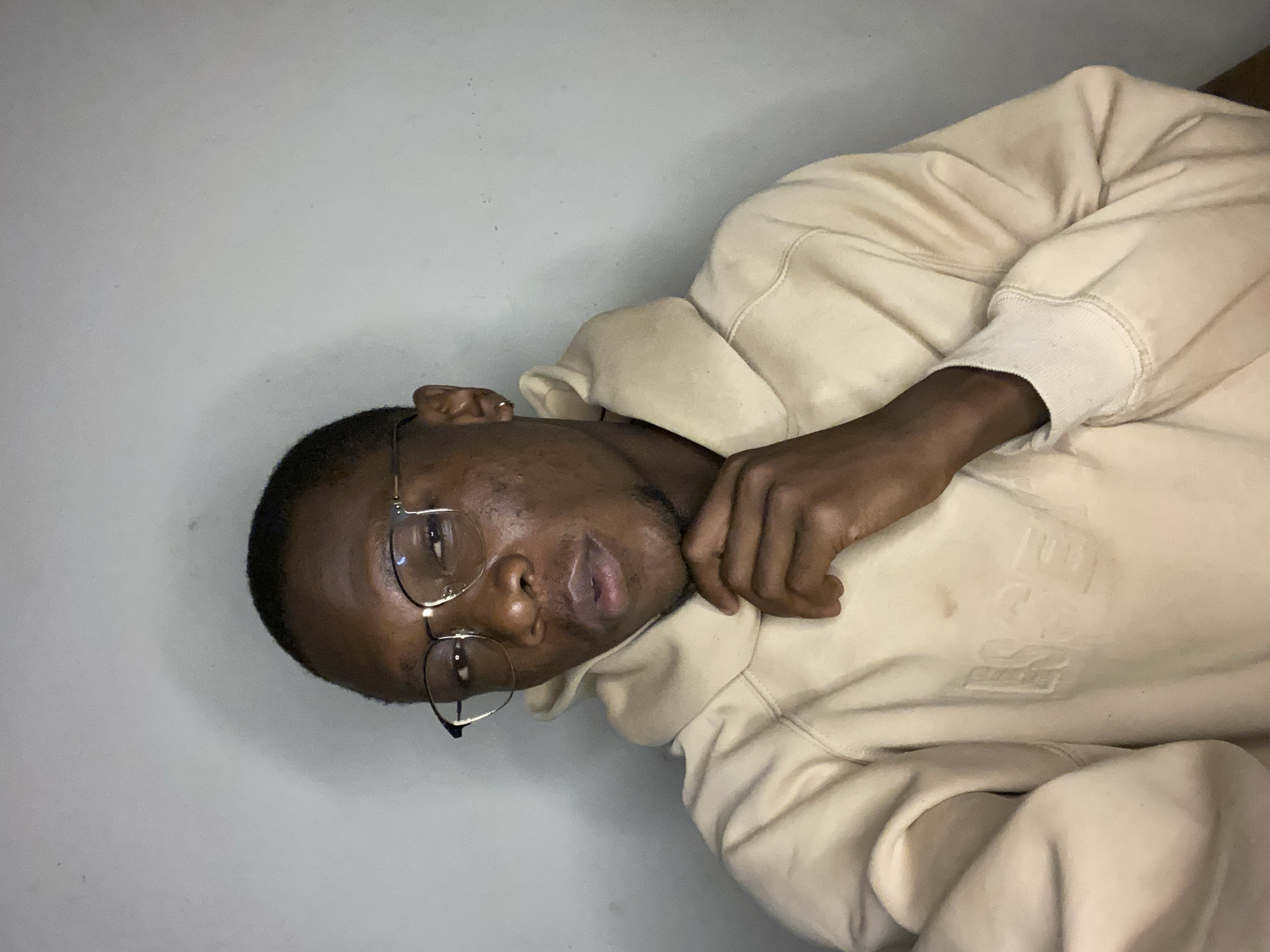 A young Black man is depicted in a close-up, horizontal portrait, appearing to be lying down and looking directly at the camera. He wears thin-rimmed glasses, which reflect some light, and has short, dark hair and light stubble on his face. His expression is neutral and calm. He is dressed in a light cream or beige-colored hooded sweatshirt (hoodie), the fabric appearing soft and thick. His left arm is bent, with his hand resting casually near his neck or cheek. On the front of the hoodie, subtle embossed lettering is visible, clearly showing "ESS", possibly part of a brand name such as "ESSENTIALS". The background is a plain, light grey wall. The lighting is soft and even, creating a relaxed and intimate atmosphere. The scene suggests an indoor setting, and the provided location context is Bukuru, Nigeria.