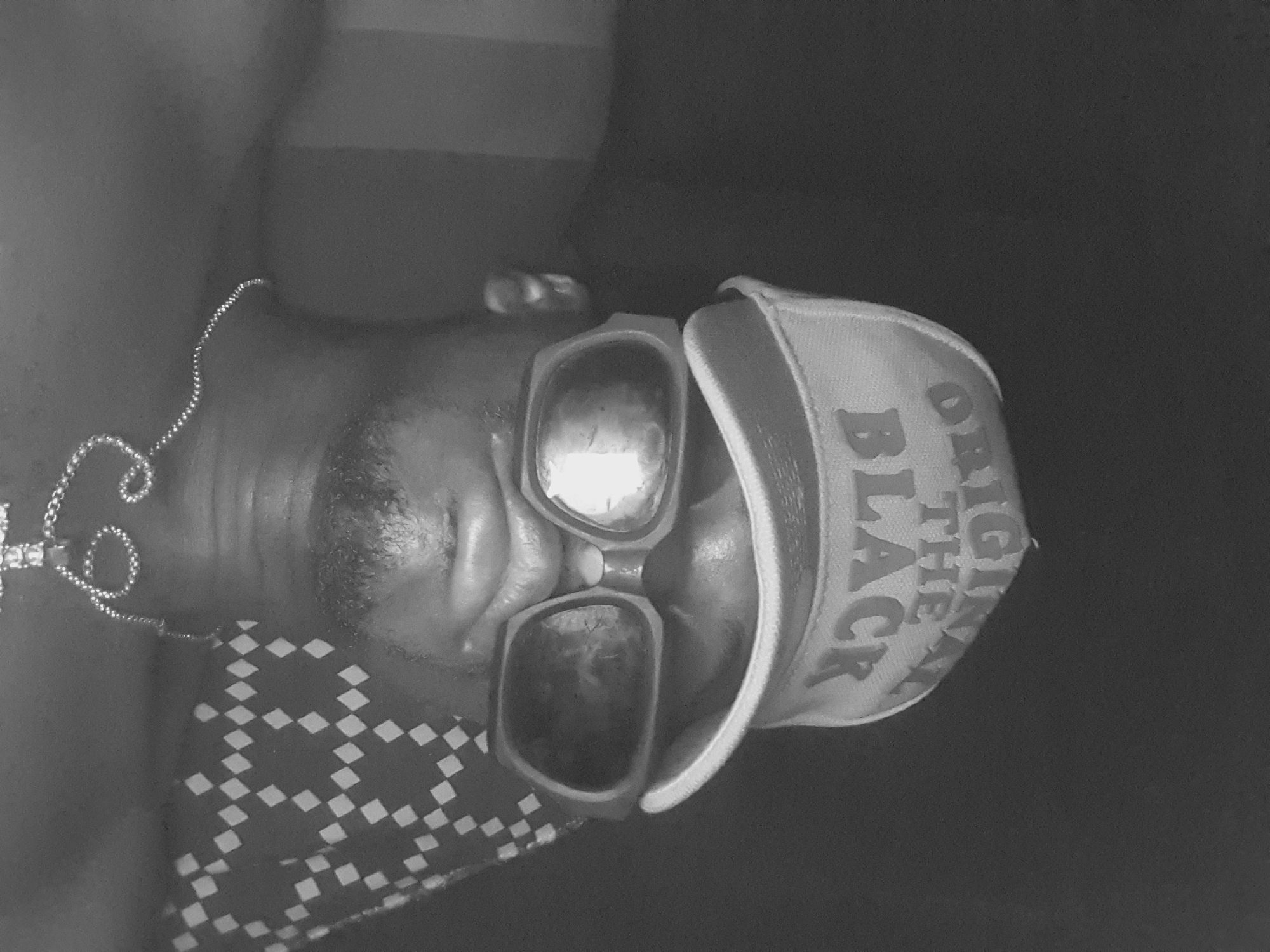 The image is a grayscale close-up of a person wearing sunglasses and a baseball cap. The text "ORIGINAL BLACK" is visible on the cap. The person is also wearing a chain necklace with a cross pendant. Part of a patterned shirt or cloth is visible on their chest. There are no indications of a location within Lokoja, Nigeria or any identifiable environmental context.