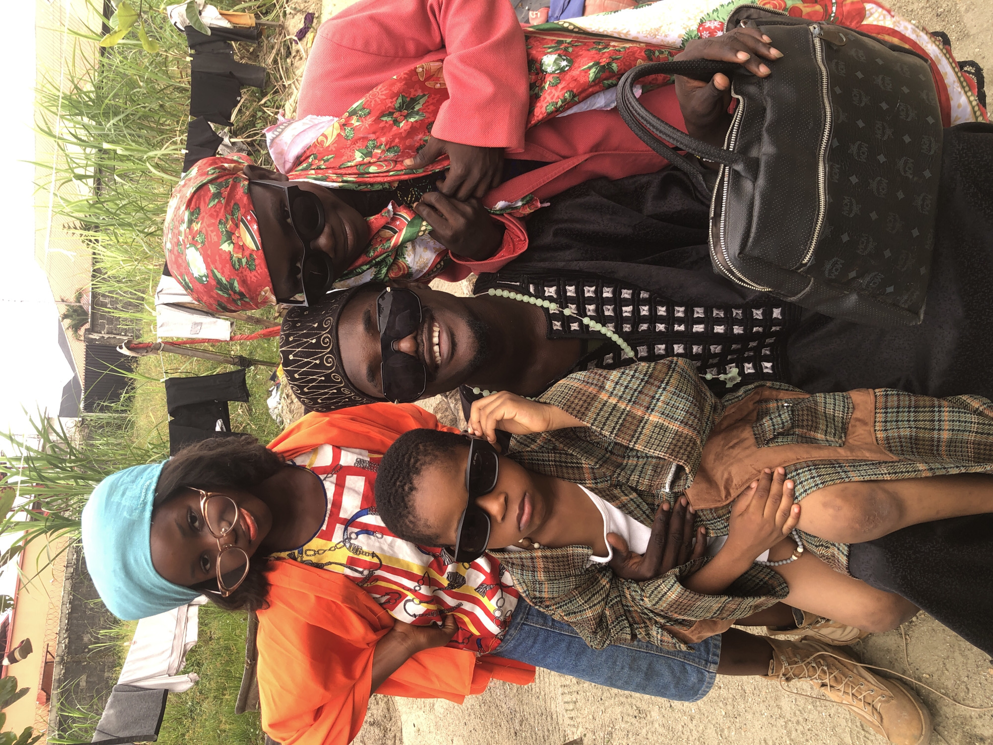 In Ado, Nigeria, four individuals are posing for a photograph outdoors. The foreground is occupied by a man in traditional attire, wearing sunglasses and a patterned cap. He is holding a young child, who is also wearing sunglasses and a plaid jacket over a white undershirt. To the man's left, a young girl in an orange jacket and blue headscarf is looking at the camera. To the man's right, another person is wearing a red jacket and a patterned headscarf, also with sunglasses. Behind the group, laundry is hanging on a clothesline, with tall green vegetation and buildings visible in the background. The ground appears to be dirt or gravel.