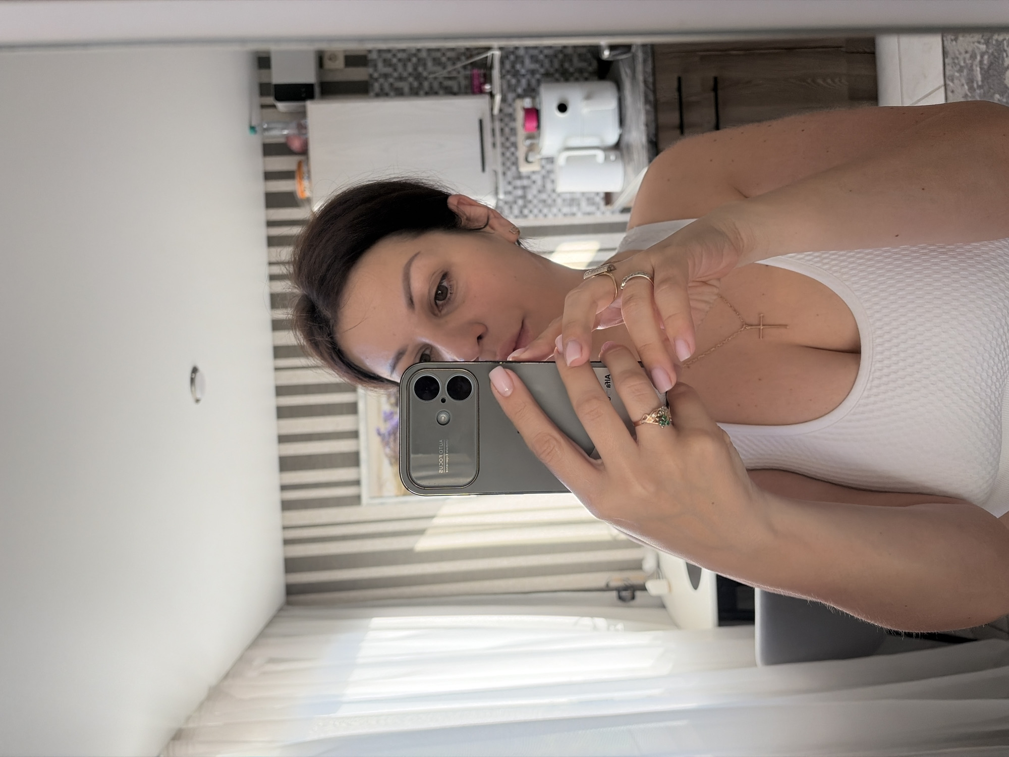 A medium shot shows a woman taking a selfie in a mirror. She is wearing a white tank top and a gold necklace with a cross pendant. She has dark hair pulled back and is wearing rings on her fingers. Her phone, with a large camera array, is visible as she holds it up to take the photo. The background appears to be a kitchen or bathroom, with cabinets, a countertop, and some appliances visible. The overall lighting is bright and natural, suggesting it is daytime.