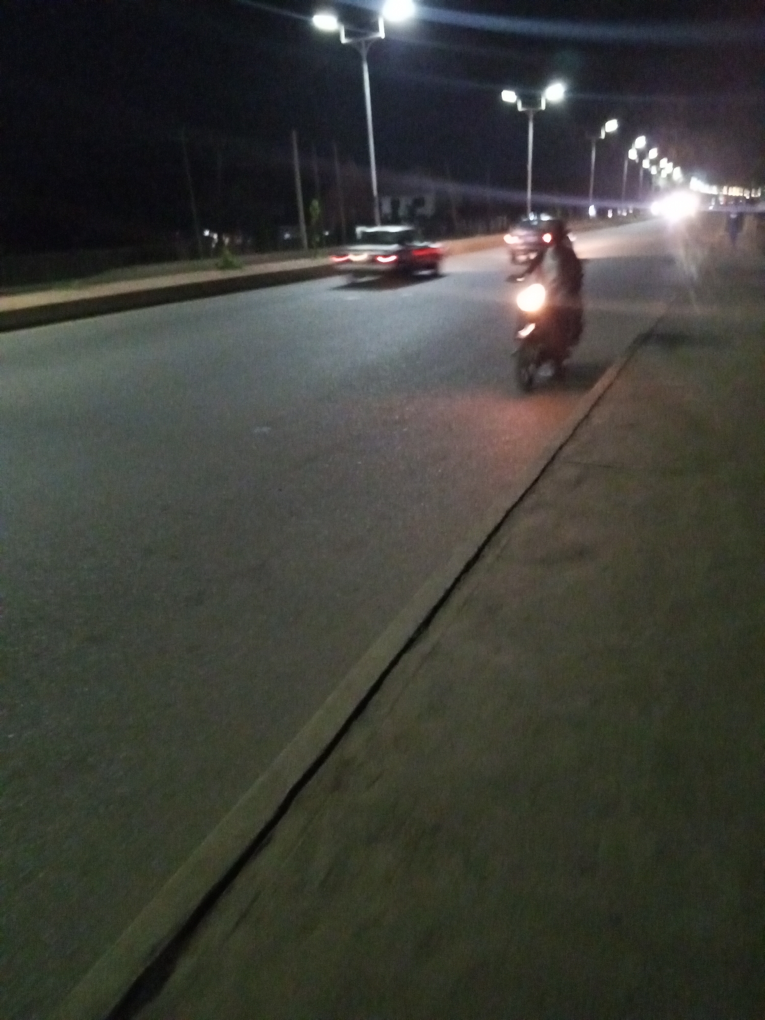 The image depicts a street scene in Bukuru, Nigeria, at night. The road is illuminated by streetlights. A motorcycle is traveling on the right side of the road, heading towards the camera. A car is also visible on the road in the distance. Other vehicles and people are seen in the distance. The environment consists of the road, sidewalks, and streetlights.
