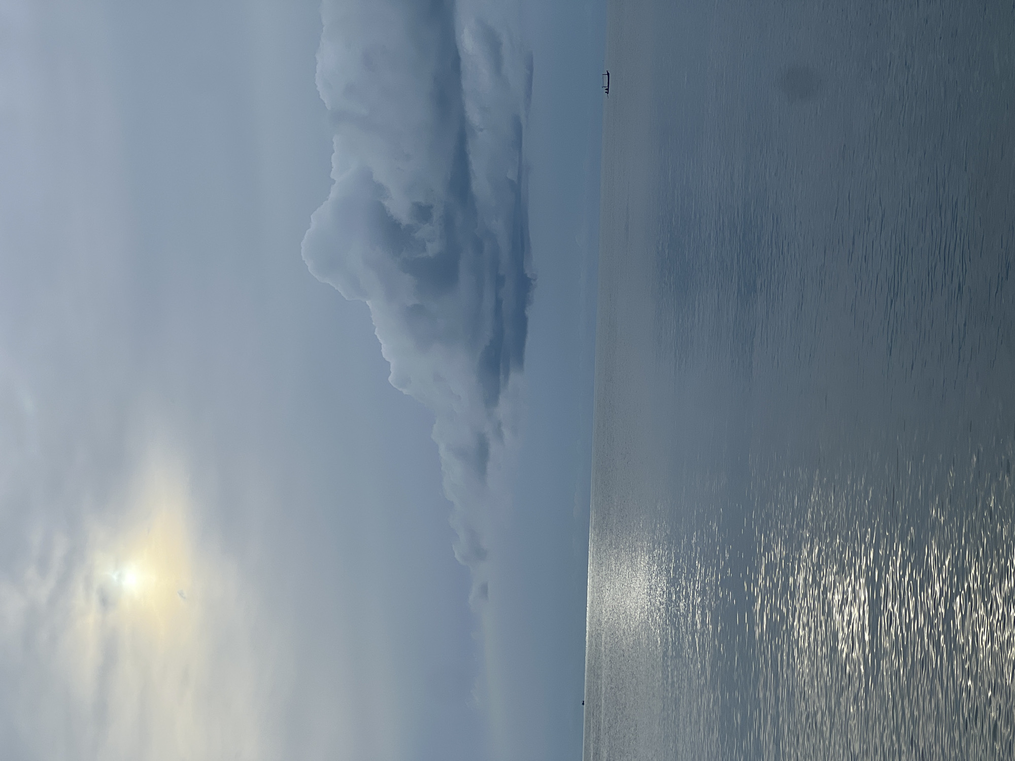 An overcast sky looms over a calm sea in Baan Chaweng Noi, Thailand. The sun, partially obscured by clouds, casts a soft, hazy glow on the upper left quadrant of the image. A large, dark cloud formation stretches across the mid-right, contrasting with the lighter clouds above. The water surface is mostly smooth, reflecting the sky, with ripples becoming more pronounced towards the foreground. Two small boats are visible in the distance. One is on the far left horizon, and the other, a narrow vessel with what appears to be two occupants, is positioned on the right horizon. The overall scene is serene and muted, suggesting a possibly cloudy or pre-storm atmosphere.
