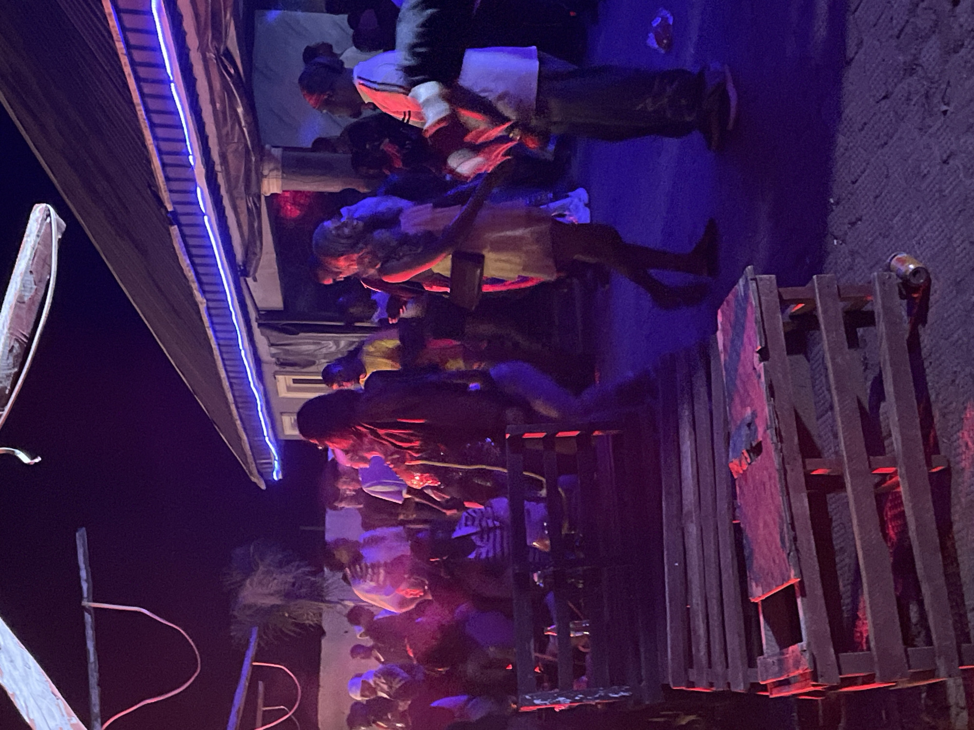The image depicts an outdoor gathering at night, possibly a party or social event, in the city of Durumi, Nigeria. A group of people are visible, some dancing and interacting. The environment appears to be illuminated by artificial lighting, with a prominent blue neon light strip running along the edge of a building's roof. Wooden pallets are arranged in the foreground, suggesting a rustic or makeshift seating area. The overall atmosphere is lively, with individuals dressed in casual attire. The ground is paved and scattered with some debris, including a discarded soda can.