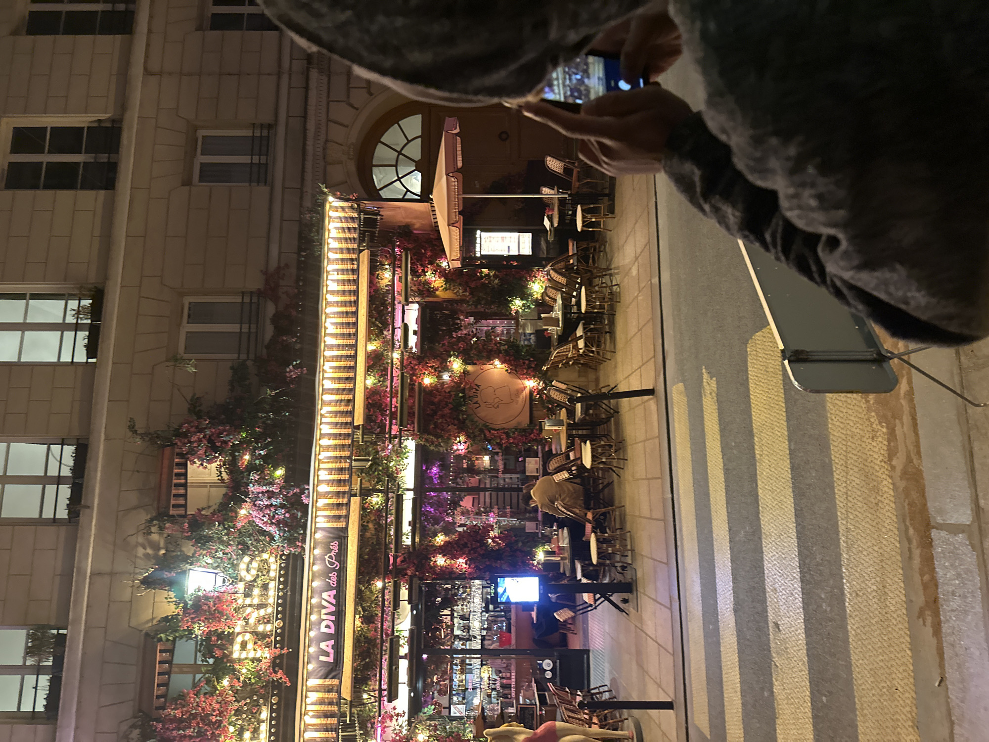 The image captures a vibrant night-time street scene in Paris, France, showing the illuminated facade of a restaurant or cafe called "LA DIVA des Près." In the foreground, a person, seen from the chest up, holds a smartphone, actively taking a picture of the lively establishment. Their head is obscured by a hood, and their right arm and hand are prominent as they operate the phone.
The cafe's exterior is heavily decorated, featuring an abundance of pink and red artificial flowers, resembling bougainvillea, draped over a trellis structure. Numerous warm-toned string lights are intertwined with the foliage and strung along the canopy, creating a bright and inviting atmosphere. Bistro-style tables and chairs are arranged on the sidewalk, suggesting an outdoor dining area. One patron is visible seated at a table, facing away from the camera. A large, folded outdoor umbrella and a digital menu display are also part of the cafe's setup.
Above the main entrance, a prominent sign or banner clearly displays the name "LA DIVA des Près." The building itself is a classic multi-story structure with light-colored stone facades and rectangular windows, some featuring decorative iron bars. An elegant arched window is visible on an upper floor. The ground before the cafe is paved, with some linear patterns that could be part of a pedestrian crossing or sidewalk design. The overall impression is one of a charming, bustling Parisian evening.