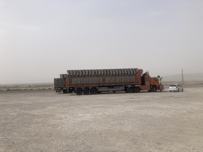 A heavily loaded semi-truck, decorated with colorful patterns, sits in a barren landscape alongside a small white car. The truck is full of cylindrical cargo. The scene is washed in hazy light, suggesting either dust or fog. A utility pole stands in the background. The ground is light-colored and appears arid.