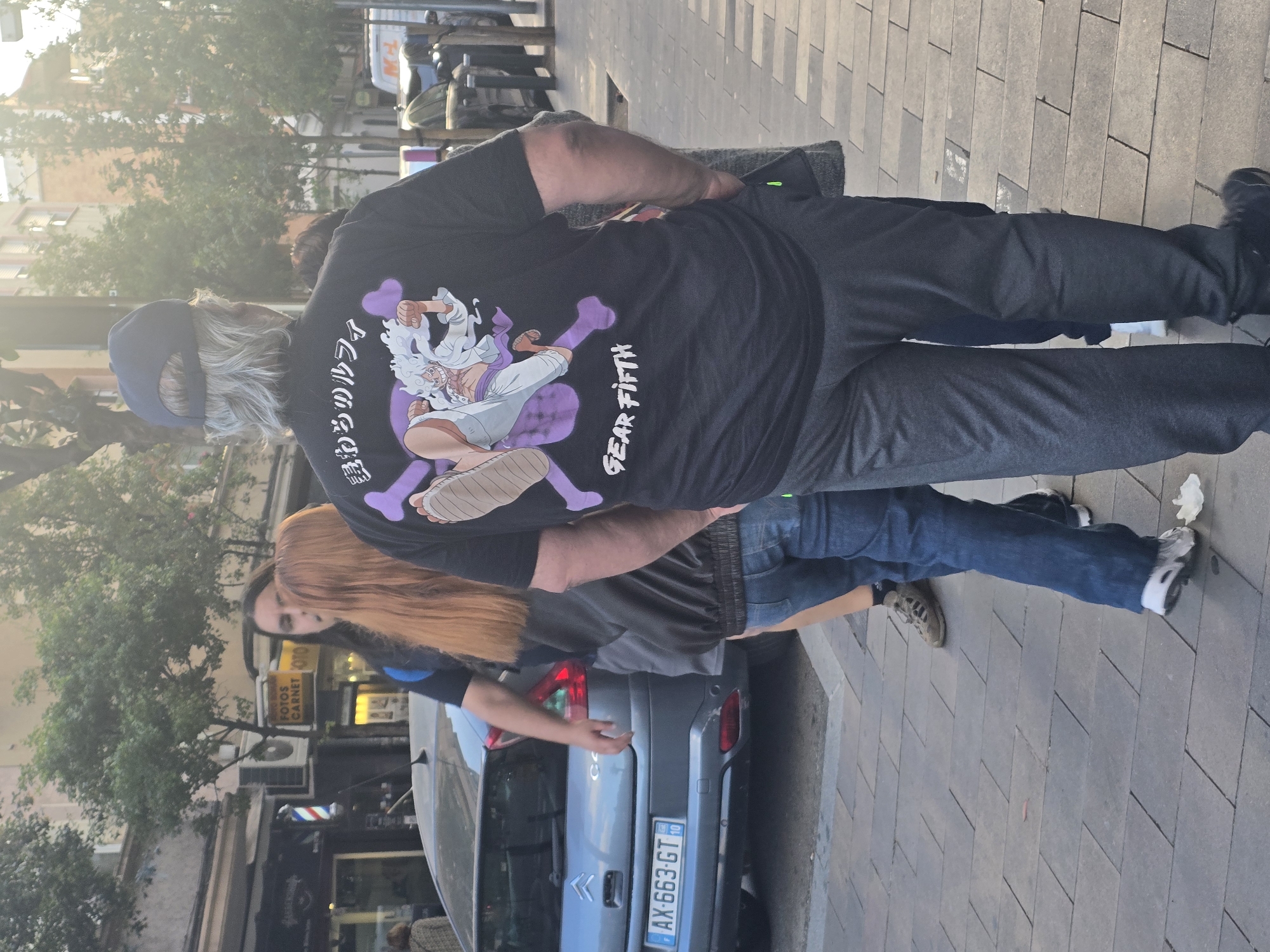 The image captures a street scene in what is likely Cornellà de Llobregat, Spain, suggested by the presence of Spanish license plates on a gray Citroen car parked along the curb. A man wearing a blue baseball cap, gray hair, a black t-shirt with a "Gear Fifth" graphic from the anime One Piece, and gray sweatpants is standing on the sidewalk. He is the most prominent subject. Beside the car, another person with long, reddish-brown hair stands facing towards the back of the car. The sidewalk is paved with gray tiles. In the background, there is a building with a sign reading "FOTOS CARNET" and a barbershop with a traditional barber pole. Trees are present on the left side of the frame.