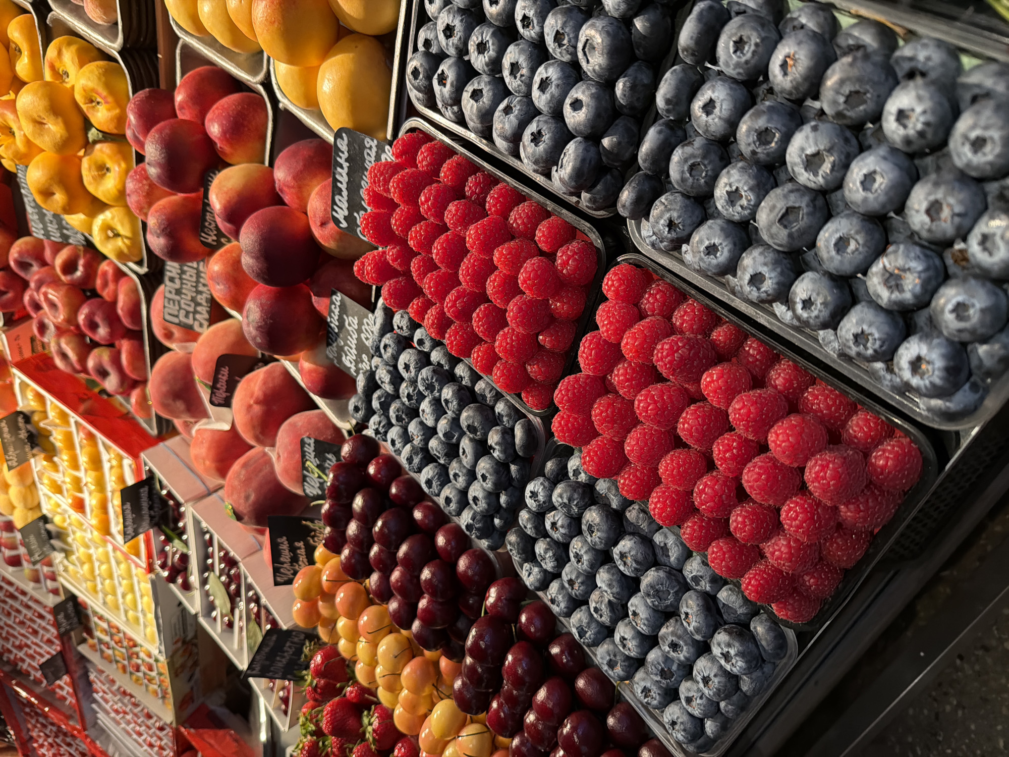 The image captures a display of fresh fruits and berries, likely at a market or grocery store. The fruits are arranged in orderly rows within plastic containers. From left to right, the fruits include yellow plums or apricots, red peaches, dark red cherries or grapes, yellow and orange plums, and red strawberries. To the right of these are blueberries and raspberries, also arranged in separate containers and rows. Small black signs with Russian writing are placed near the fruit, likely indicating prices or types. The location is Moscow, Russia. The scene suggests a well-stocked fruit section with an emphasis on visual appeal.