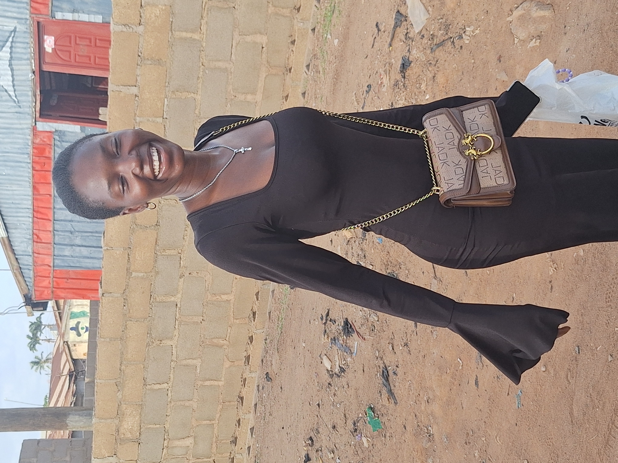 A smiling young woman with short hair stands outdoors in Sabon Tasha, Nigeria. She wears a long-sleeved black dress and a silver necklace with a cross pendant. A brown purse with a gold chain is slung over her shoulder. She is standing on sandy ground littered with debris and plastic. Behind her is a building made of cinder blocks with a red-framed window and a corrugated metal roof. Palm trees are visible in the distance.
