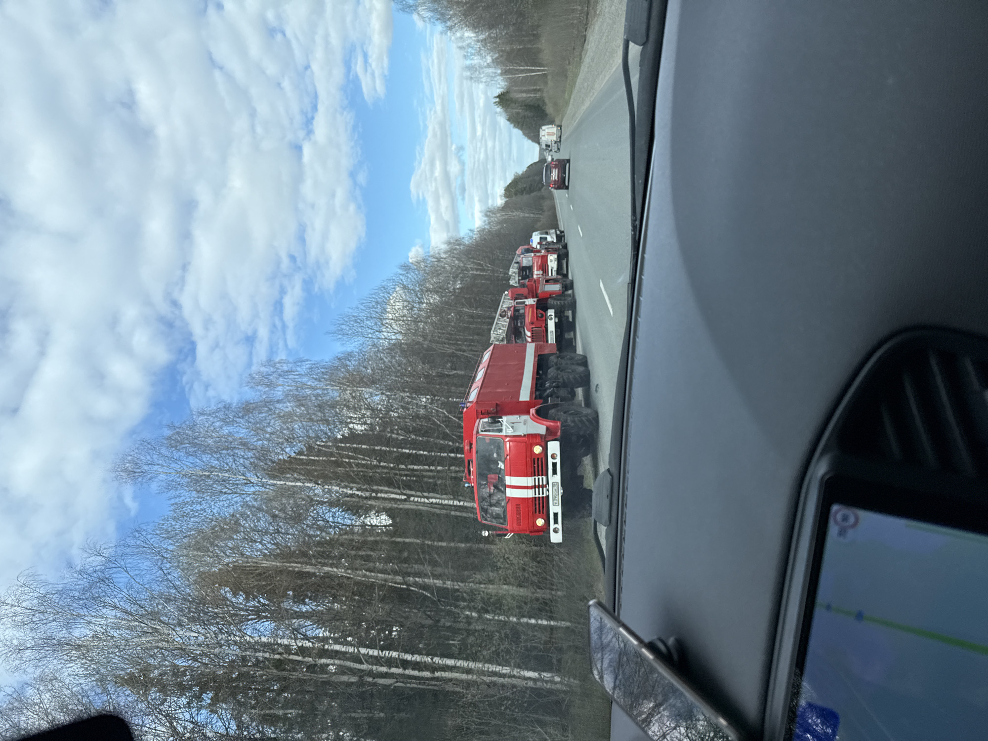 The image shows a line of red and white fire trucks driving on a highway, as seen from inside a vehicle. Bare trees line the left side of the road, while other vehicles are visible in the distance. The sky is partly cloudy. A navigation screen is visible on the dashboard of the vehicle from which the photo was taken. The location is Iiul'skoe, Russia.