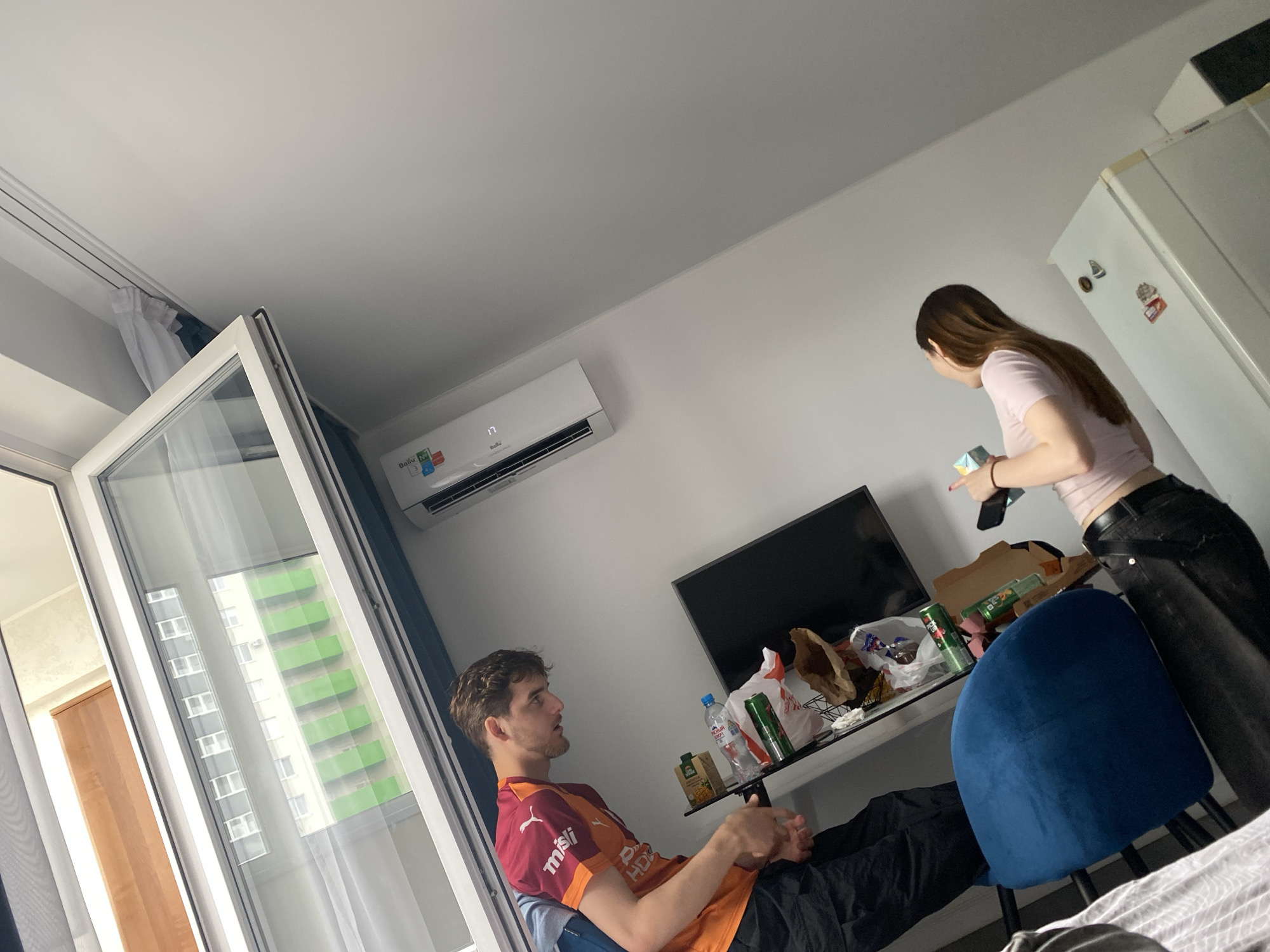 In a room with white walls and a white ceiling, a man and a woman are present. The man, wearing a maroon and orange shirt, is seated on a blue chair, looking to his right. The woman, with long brown hair and wearing a pink shirt and black pants, stands near a white refrigerator, looking down at a box and a phone in her hands. On a table between them are various items including a television, drinks, and food packaging. The room appears to be a living space, possibly an apartment in Ufa, Russia, with a window on the left revealing a view of a green building.