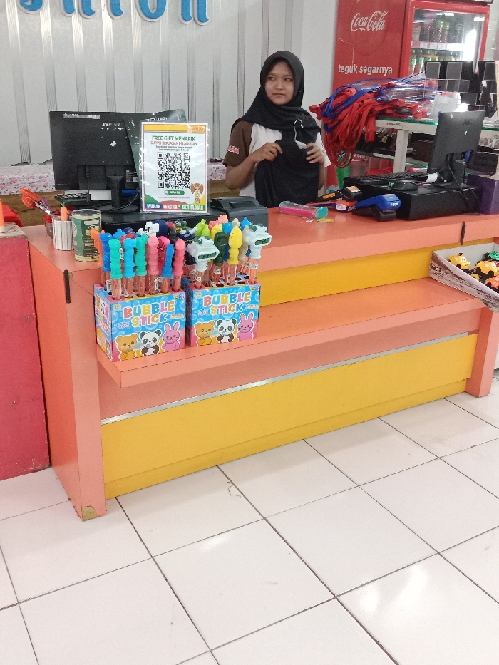 This image depicts an indoor retail environment located in Langenharjo, Indonesia. A young adult female, wearing a black hijab and a light-colored polo shirt with a brown collar, stands behind a multi-tiered counter. She holds a black garment in both hands, positioned at waist level, and looks towards the viewer's left with a neutral expression. The counter is constructed with orange and yellow panels. On the left side of the counter, two cardboard display boxes labeled "BUBBLE STICK" with cartoon graphics contain multiple colorful bubble wands. Behind these, a black computer monitor and keyboard are visible. Centrally positioned, a white vertical sign displays a QR code and Indonesian text offering a "FREE GIFT" and mentioning a customer satisfaction survey. To the right of the individual, a second black computer monitor, a blue payment terminal, and a black cash register are present. A small open box containing multiple miniature toy vehicles is visible on a lower counter shelf to the right. The background features a white paneled wall with large blue lettering partially visible at the top. A red Coca-Cola branded refrigerator with Indonesian text ("teguk segarnya") is situated on the right, next to a white shelving unit stacked with folded red garments. The floor consists of white square tiles with dark grout lines.