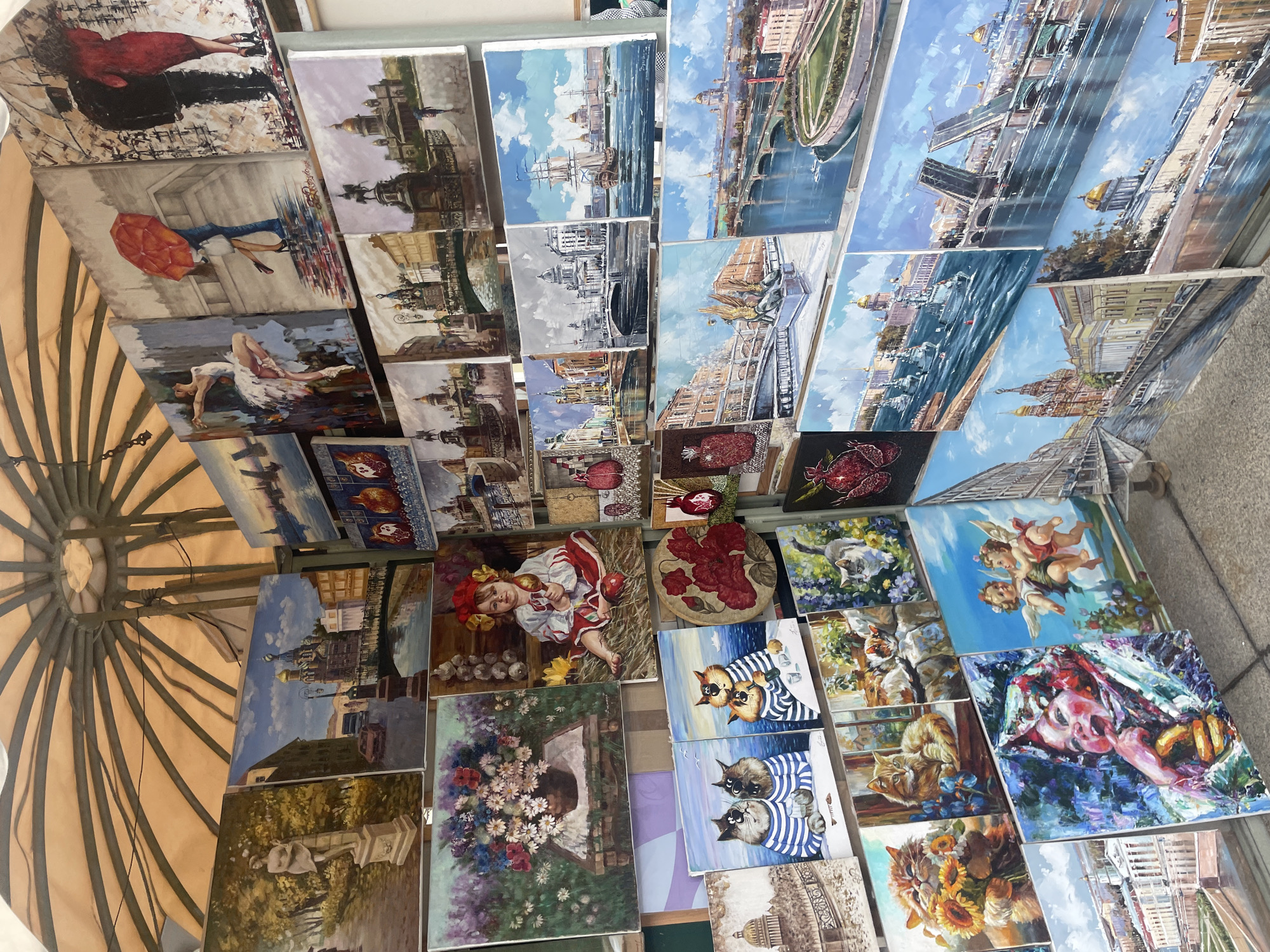 This image depicts an outdoor art stall in Saint Petersburg, Russia, displaying a variety of oil paintings. The stall is set up under a large, round, beige awning supported by metal poles. The paintings are arranged in multiple rows, leaning against each other and supported by stands. The subjects of the paintings include cityscapes of Saint Petersburg, featuring iconic landmarks such as cathedrals with golden domes, bridges over canals, and historic architecture. There are also genre paintings, including depictions of couples under umbrellas, a ballerina, a child in traditional attire, still lifes of flowers, and playful portraits of cats. The ground is paved with cobblestones. The lighting suggests it is daytime, and the overall scene conveys a vibrant artistic atmosphere.