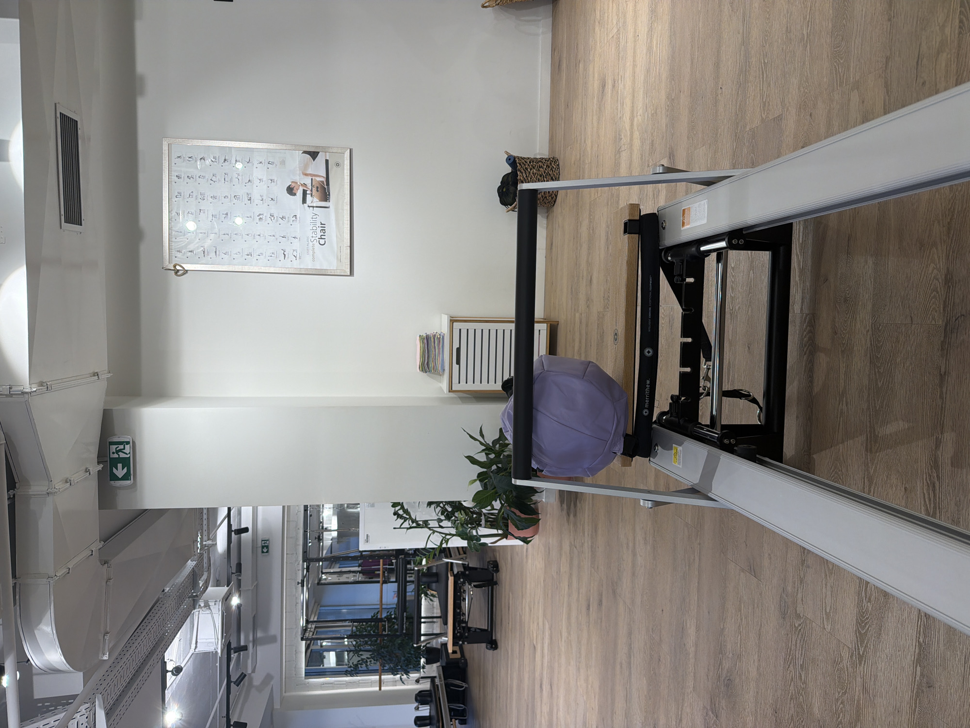 The image shows the interior of a Pilates studio. In the foreground, a Pilates reformer machine is visible, with its metal rails and wooden seat. A light purple exercise ball rests on the seat. In the background, other Pilates equipment, such as reformers and Cadillac tables, are set up. Large windows in the back reveal a glimpse of what appears to be an urban environment, possibly including buildings and streetlights. The walls are white, and the floor is made of light-colored wood. There are also plants and storage units in the studio. On the wall, a framed chart displays various Pilates exercises. Two green exit signs with running figures are also visible on the wall and ceiling. The overall impression is of a clean, modern, and well-equipped fitness facility. The location is described as Boreti, Montenegro.