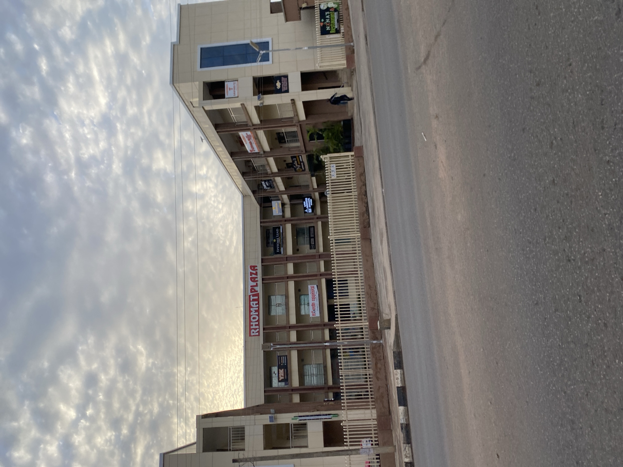 The image shows a three-story building with the prominent sign "RHOMAT PLAZA" in red lettering. The building is light beige with brown accents and has multiple windows and balconies. Various smaller signs for businesses are visible on the facade, including "Y2K", "EPITOME LEAD", "Rosa's apparel", "Fitness-NB", and "NXNA's Restaurant". A white metal fence encloses the front of the property. A single person in dark clothing is walking towards the right side of the building near the entrance. The building is situated on a street with a paved road and a sidewalk with a black and white checkered curb. Power lines are visible against a cloudy sky. The location is identified as Bar Jirgi Summa, Nigeria.