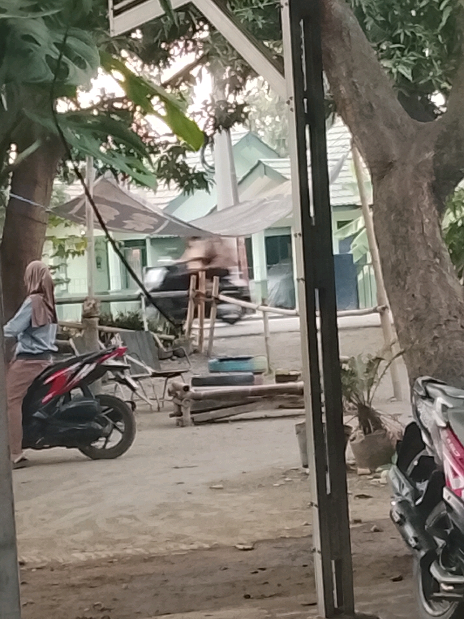 The image depicts an outdoor scene, likely in Ujungrusi, Indonesia, characterized by a sandy or dirt ground surface. In the foreground, a red and black motorcycle is parked. To the left, a person wearing a light blue long-sleeved shirt and a brown hijab is standing and facing away from the camera. Behind the motorcycle and the person, a low-lying structure with a light-colored roof and green trim is visible. A large tree with a rough bark trunk is situated on the right side of the frame. The background reveals more trees and vegetation, suggesting a semi-rural or suburban environment. The lighting indicates it is daytime, possibly late afternoon, with sunlight filtering through the trees.
