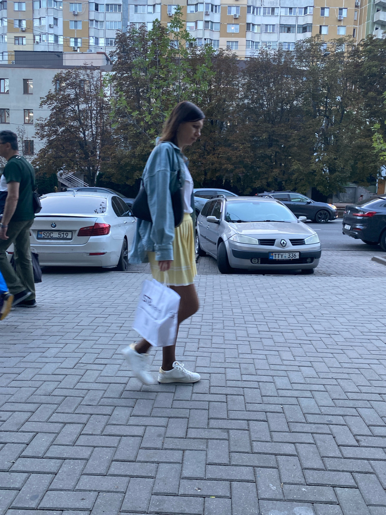 The image depicts a street scene in Chișinău, Moldova. In the foreground, a young woman is walking from left to right across a brick-paved sidewalk, carrying a white shopping bag. She is wearing a light blue denim jacket over a white t-shirt and a yellow skirt, with white sneakers. Behind her, to the left, a person in a green t-shirt and dark pants is walking away from the camera. Several cars are parked on the street, including a white BMW with license plate "SQC 519" and a silver Renault station wagon with license plate "TTV 336". In the background, there are multi-story apartment buildings and trees with autumn foliage. The overall impression is that of a casual urban daytime scene.