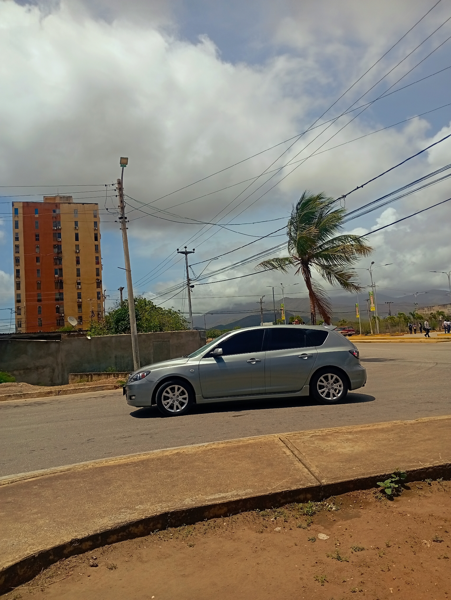 A light silver hatchback car is driving on a paved road in a city. The car is in motion, as indicated by the slight blur of the wheels. To the left of the car, there is a tall, narrow apartment building with orange and yellow siding, featuring many windows and air conditioning units. A gray concrete wall runs in front of the building. A street lamp is visible on a utility pole near the car. Further in the background, there is a palm tree with its fronds being blown by the wind. Beyond the palm tree, mountains are visible under a cloudy sky with some patches of blue. Several utility poles with numerous wires are strung across the scene. In the distance, to the right, there are other buildings and people walking, suggesting an urban or semi-urban environment. The road has a concrete curb and a sandy, dirt shoulder with some sparse vegetation. The scene appears to be captured during daylight, with the sun casting shadows.