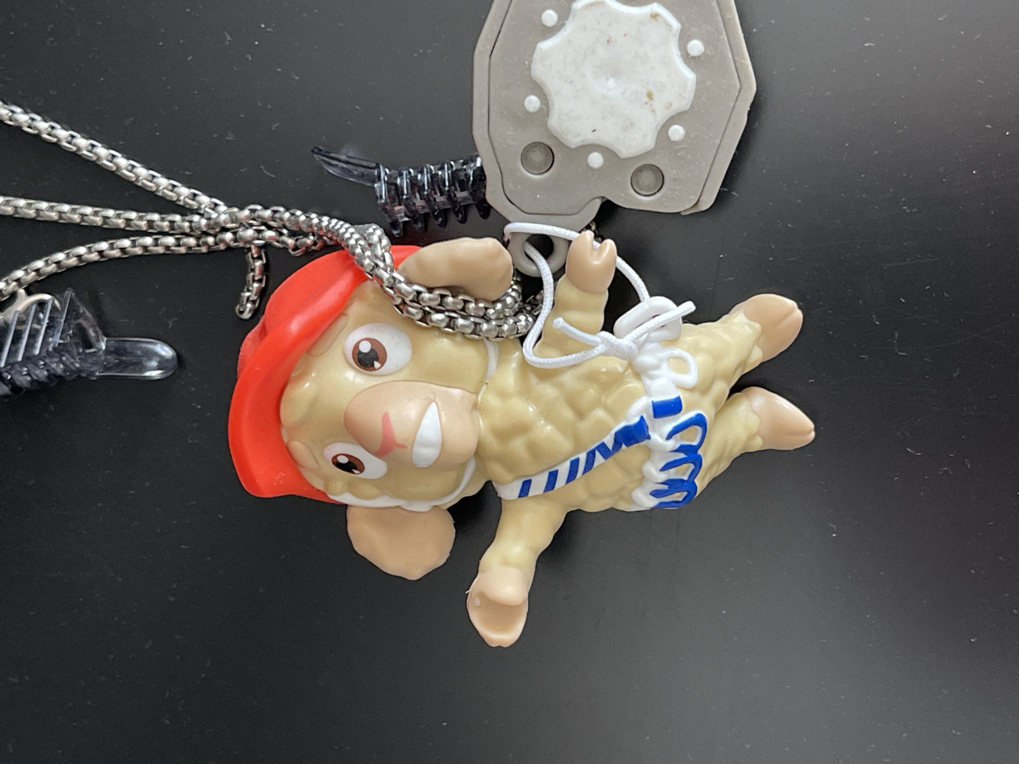 A close-up, overhead shot shows a toy sheep figure with a red hat and a blue and white sash, possibly a character from an anime or cartoon. The figure is attached to a silver chain necklace. To the right of the figure is a gray, octagonal object with a white inset, resembling a dog tag or a medallion. The background is a dark, textured surface, likely a table. The image composition is simple, with the focus solely on the toy and its accessories. There is no discernible connection to Kyiv, Ukraine, nor any indication of people or actions.
