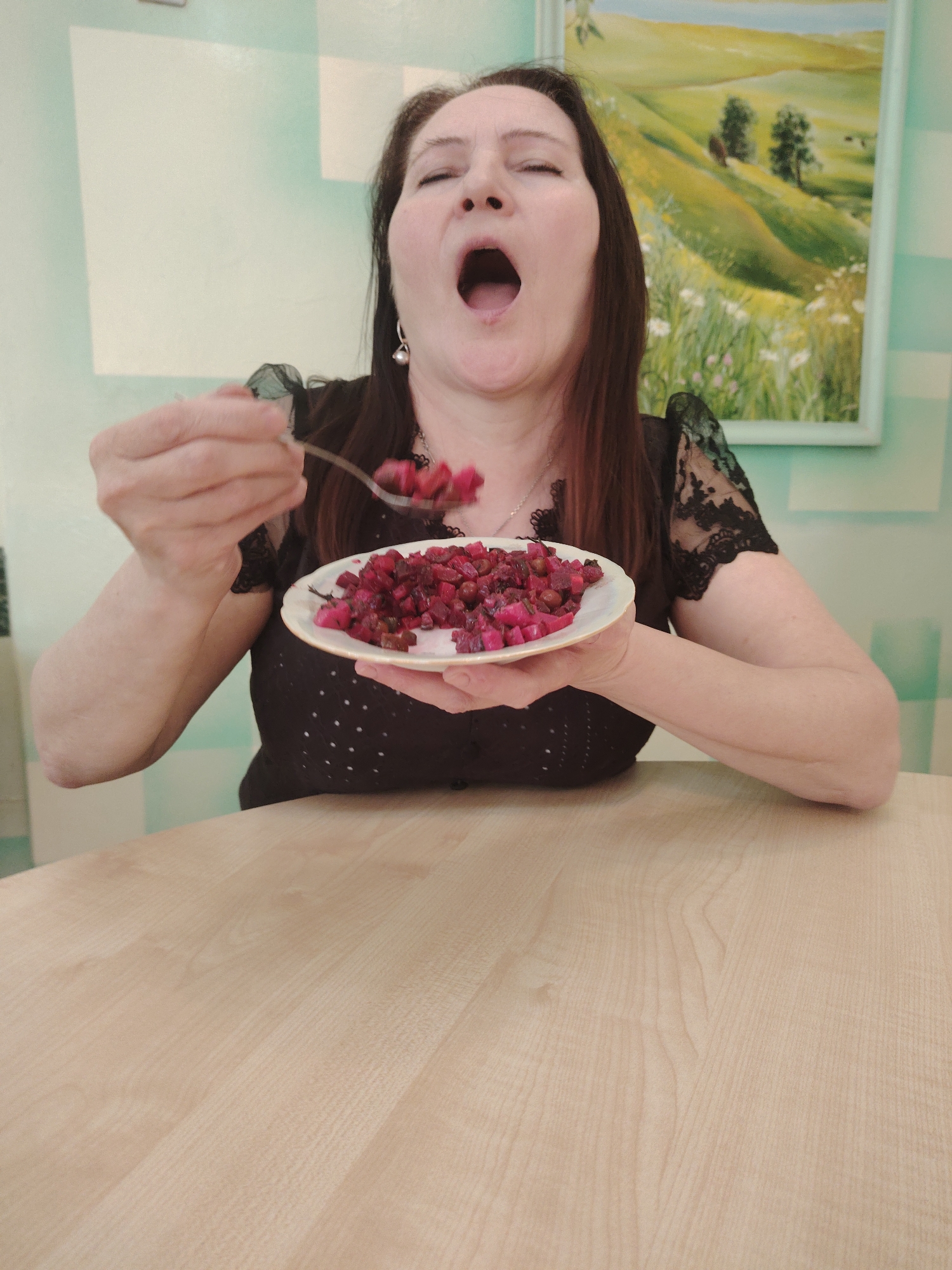 A middle-aged woman with dark hair and an open mouth is seated at a light-colored wooden table, holding a plate of beetroot salad (vinegret) in her left hand and a fork with the same salad in her right hand, in front of her open mouth. She is wearing a black blouse with lace sleeves and a necklace. Behind her is a wall with light green and white stripes, and a framed landscape painting. The location appears to be inside a building, possibly a restaurant or cafe, identified to be in Sarapul, Russia.