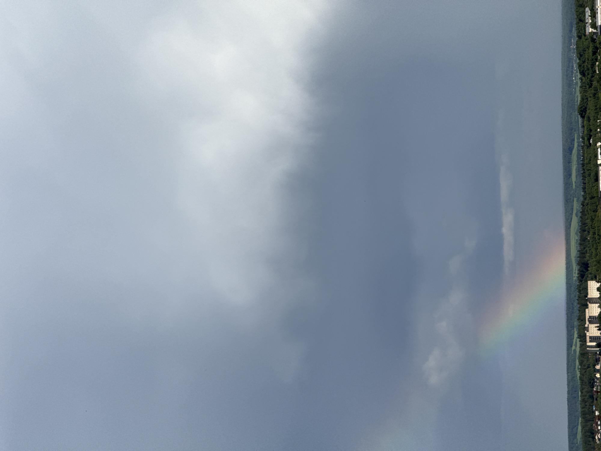 The image shows a cityscape under a cloudy sky with a rainbow on the right side of the frame. The city appears to be located in Unknown, Unknown. A green belt of trees is visible separating the urban area from the sky. Buildings are seen along the edge of the city where the rainbow touches down. The sky is predominantly gray and white, indicating overcast conditions.