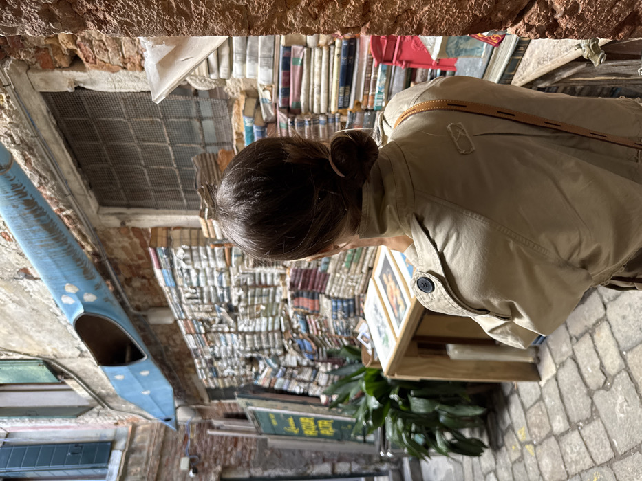 The image captures a narrow, brick-walled alleyway within the Libreria Acqua Alta in Venice, Italy. A person, viewed from behind, with dark brown hair tied in a bun, is positioned centrally. They wear a light tan trench coat, with a visible brown leather strap over their right shoulder. The person is standing before a wooden display stand holding two framed artworks or prints. To the left of this stand, a tall green potted plant is visible. The immediate background features extensive, densely stacked old books, some forming a wall-like structure. A window with a metal security grate is centered within the brick wall above the books. A distinct blue, truncated gondola is suspended horizontally overhead. To the far left, a green sign partially displays the text "Acqua Alta." The ground is paved with weathered cobblestones. A red metal object, possibly a mailbox, is partially visible on the right side, integrated with more book stacks.