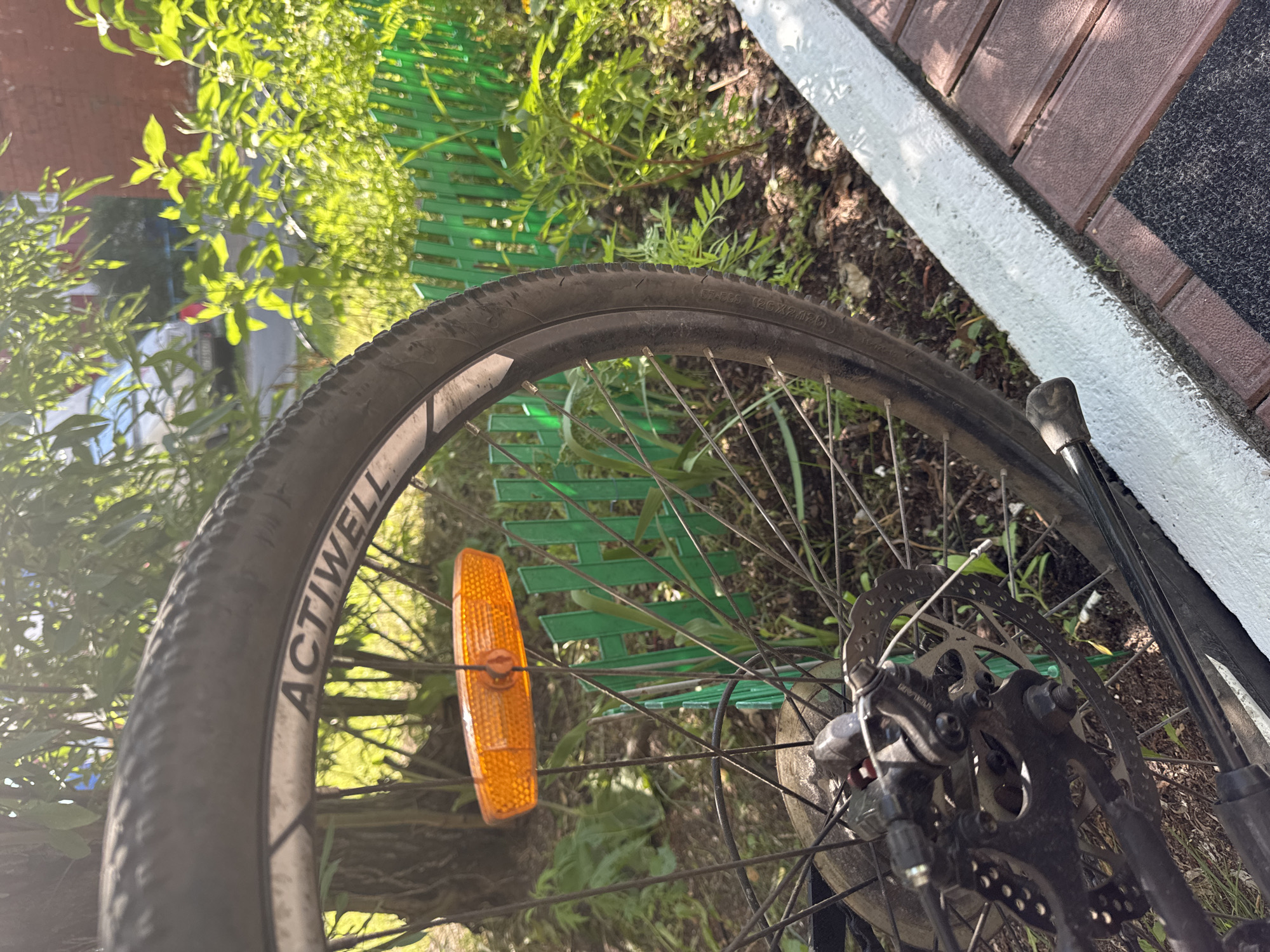 The image shows the rear wheel of a bicycle with a reflective orange spoke reflector. The bicycle is positioned outdoors, with a blurred background of green foliage, a light green fence, and a brick building. The foreground consists of a white curb and reddish-brown paving stones. The scene appears to be in an urban environment. The text on the tire reads "ACTIVELL 126X2.0(47)". The image was taken in Surgut, Russia. There are no people or animals visible in the image.