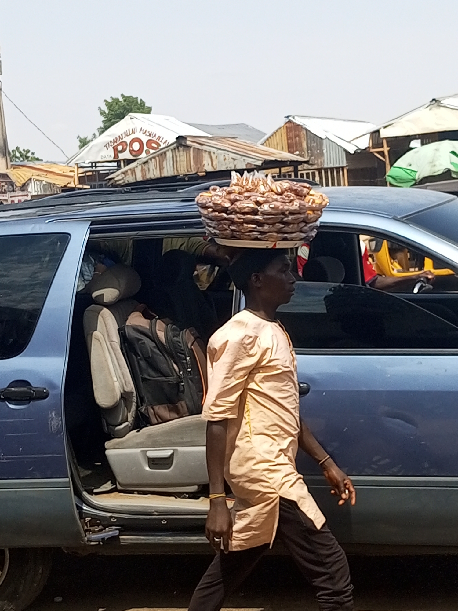 The photograph captures a vibrant street scene, likely in a market area during bright daylight. The primary subject is a dark-skinned person, seen in profile, walking from left to right while expertly balancing a large, flat tray of reddish-brown items on their head. These items appear to be numerous small, uniformly shaped snacks, possibly local confectionery or dried fruit, stacked in layers. The person wears a light-colored, long-sleeved tunic (possibly a kaftan) and dark trousers. A light-colored band is visible on their left wrist.
In the mid-ground, a blue minivan or SUV dominates much of the frame. Its side door is open, revealing the car's interior, including a light-colored seat and a backpack. The vehicle shows some signs of wear. Through the windows of the blue vehicle, a partially visible yellow vehicle and a driver's arm can be seen, indicating ongoing traffic.
The background features several informal structures with corrugated metal roofs and wooden supports, characteristic of market stalls or small roadside businesses. One notable structure has a white tarp covering its roof. Visible text on this tarp reads "TABARAKALLAH MASHAA ALLAH" (an Arabic phrase) and prominently "DATA POS" in red, with "DATA" written smaller. These structures suggest a commercial environment.
The scene depicts a common sight of street hawking and daily commerce in a bustling urban or semi-urban setting. The bright, clear lighting suggests it is sunny, likely midday or early afternoon. The activity of the person carrying goods on their head, combined with the roadside stalls and vehicles, paints a picture of everyday life in Gombe, Nigeria, where such informal trade is prevalent.