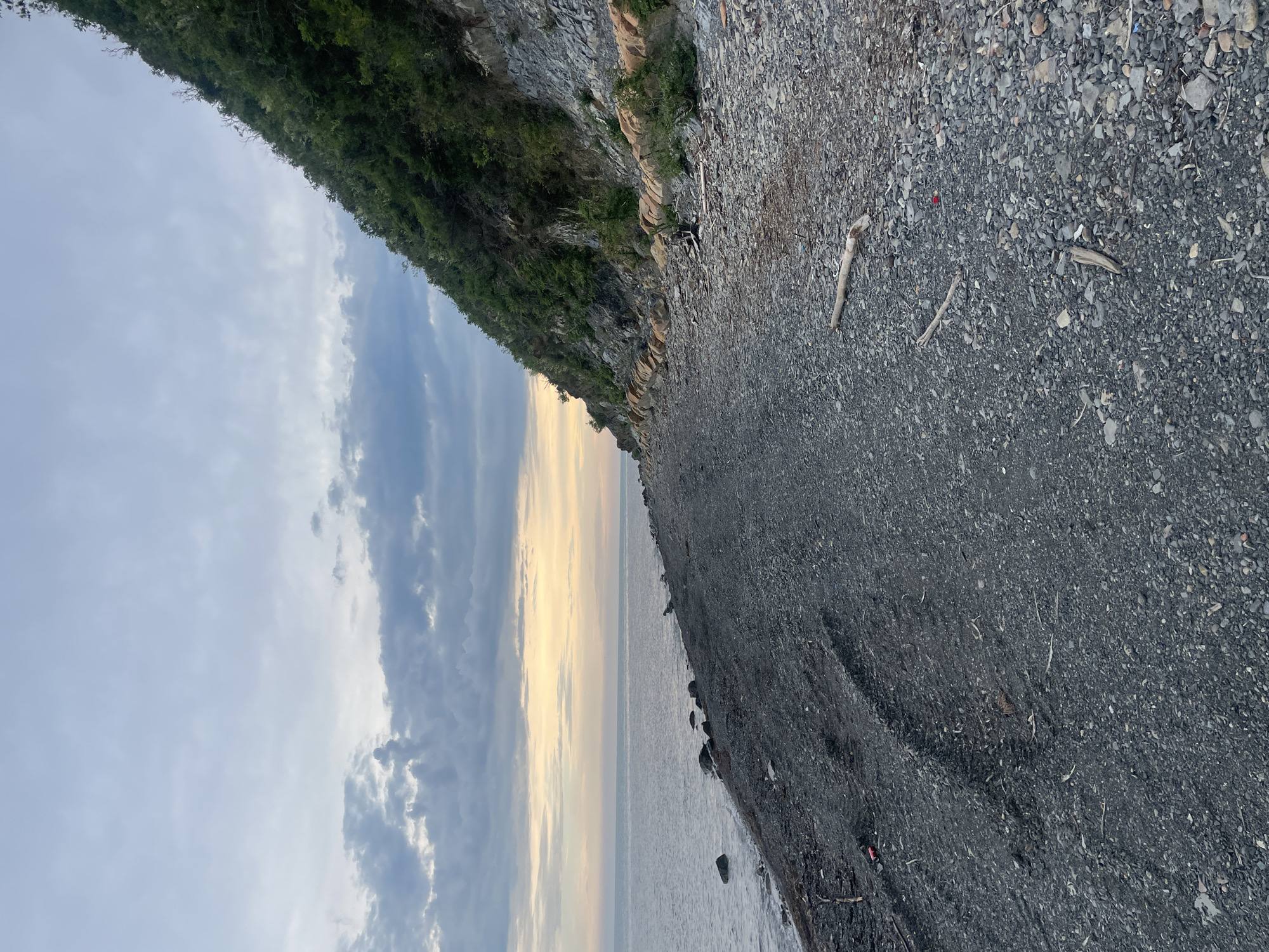The image depicts a rocky beach in Olginka, Russia, adjacent to a cliff face covered with vegetation. The beach is composed of small, dark-colored pebbles and some driftwood. The water line is visible, and the sky is filled with clouds and a pale, setting sun or rising sun. No people are visible in the frame.