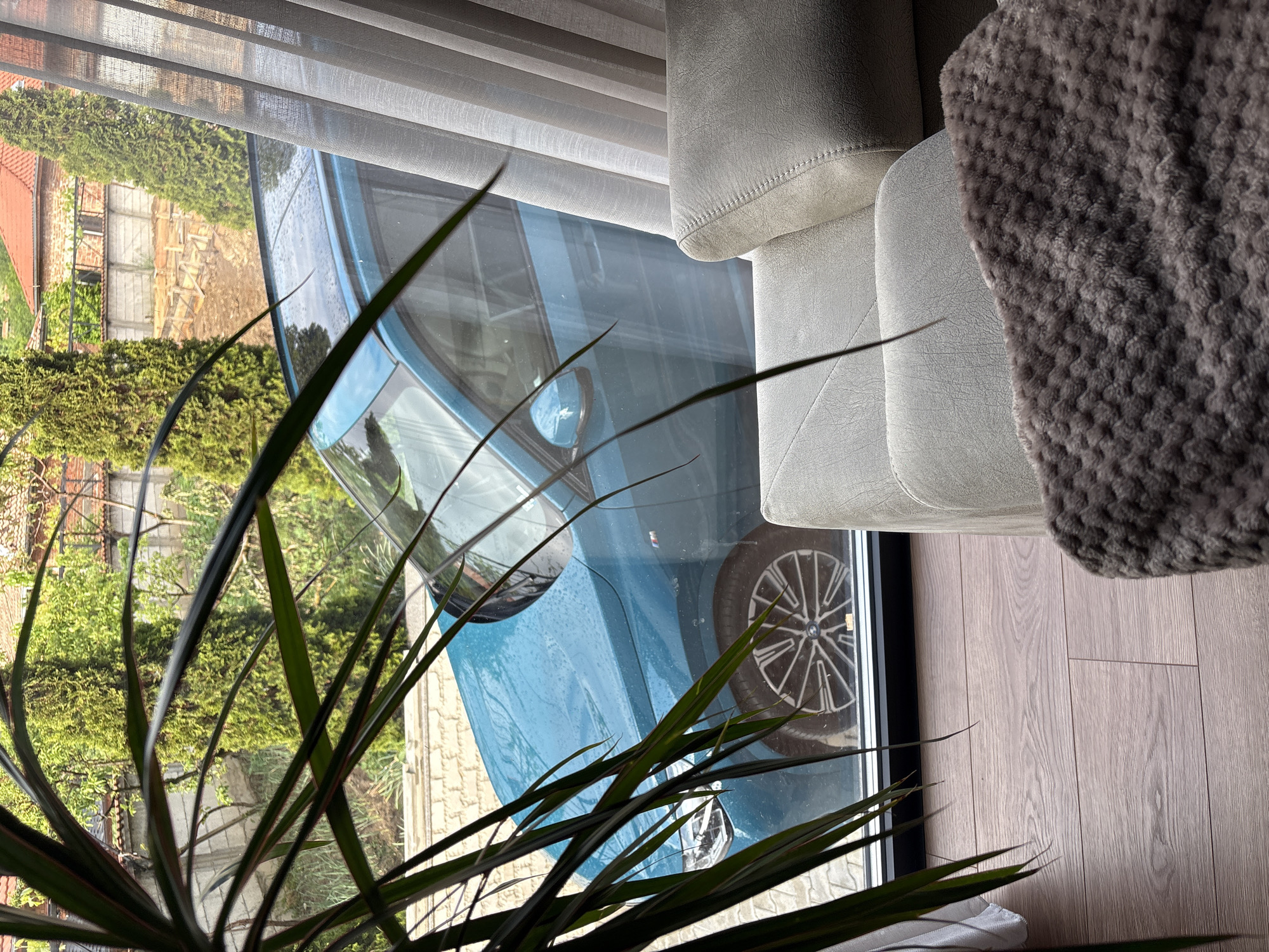 The image shows an interior view with a focus on a window reflection. A blue car is visible through the window, reflected among the interior elements. A white couch with a gray throw blanket is partially visible on the right side of the frame. Green plants are present in the lower-left corner. The view outside the window reveals parts of a building, trees, and possibly a brick walkway. The location is Dolno Drianovo, Bulgaria.