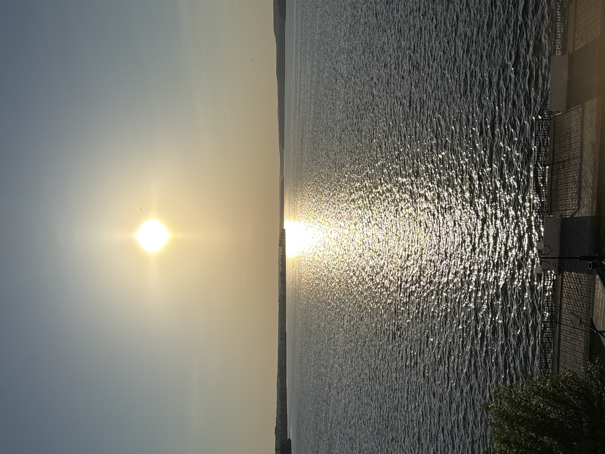 The image depicts a serene scene of the Votkinsk reservoir at sunset in Votkinsk, Russia. The sky is a gradient of blue and gold, with the sun positioned centrally and slightly to the left. The reservoir's surface is rippled, reflecting the sunlight in a shimmering effect. The opposite shore, lightly forested, is visible in the distance. A man-made embankment with a railing, lighting posts, and some greenery is seen in the foreground on the right.