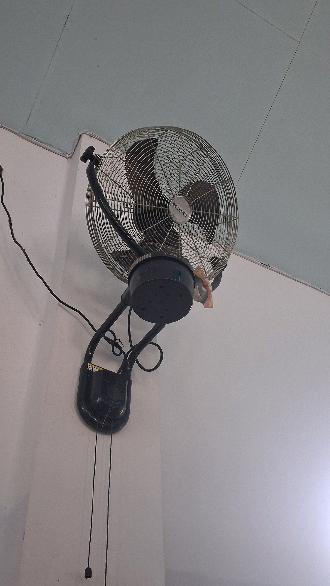 An oscillating fan is mounted on a white wall, angled upwards towards a light blue ceiling. The fan, made of chrome and black plastic, has a wire grille enclosing its blades and a visible "REGENCY TORNADO" logo on its motor housing. A light brown, folded piece of paper is tucked into the fan grille. Two black pull cords hang from the fan's mounting bracket, one of which has a dark gray toggle at its end. A black electrical cord extends from the fan to the left, following the wall and disappearing towards the upper left corner of the frame. The wall has a textured finish, and the ceiling appears to be made of large panels with visible seams. This scene is located in Giriwarno, Indonesia.