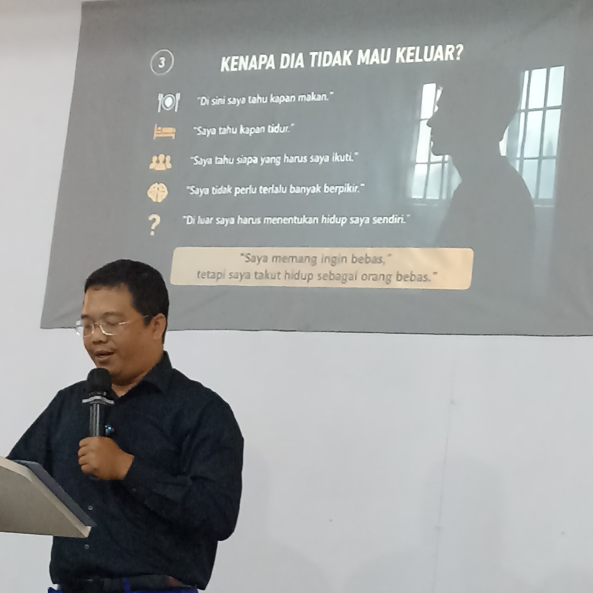 This is a medium shot, eye-level photograph taken indoors, likely in the city of Magelang, Indonesia. A man wearing glasses and a dark blue collared shirt stands on the left, holding a microphone in his right hand and a book or tablet in his left. He appears to be speaking to an audience, though no audience members are visible in the frame.
Behind the man, a large projection screen displays text and an image. The text at the top of the screen reads, "KENAPA DIA TIDAK MAU KELUAR?" which translates to "WHY DOES HE NOT WANT TO GO OUT?". Below this title, a list of bullet points with accompanying icons is displayed:
* An icon of cutlery with the text, "Di sini saya tahu kapan makan." ("Here I know when to eat.")
* An icon of a bed with the text, "Saya tahu kapan tidur." ("I know when to sleep.")
* An icon of a group of people with the text, "Saya tahu siapa yang harus saya ikuti." ("I know who to follow.")
* An icon of a brain with the text, "Saya tidak perlu terlalu banyak berpikir." ("I don't need to think too much.")
* A question mark icon with the text, "Di luar saya harus menentukan hidup saya sendiri." ("Outside I have to determine my own life.")
Below these bullet points, there is a highlighted section in a pale yellow rectangle with the text:
"Saya memang ingin bebas," ("I do want to be free,")
"tetapi saya takut hidup sebagai orang bebas." ("but I am afraid to live as a free person.")
To the right of the text, a silhouette of a person's head and shoulders is projected onto the screen, seen in profile facing left. Behind the silhouette, the projection shows what appears to be a window with multiple panes, suggesting an indoor setting with a view to the outside.
The lighting in the room appears to be artificial and moderately bright. The overall tone of the image is informative, possibly part of a presentation or seminar.